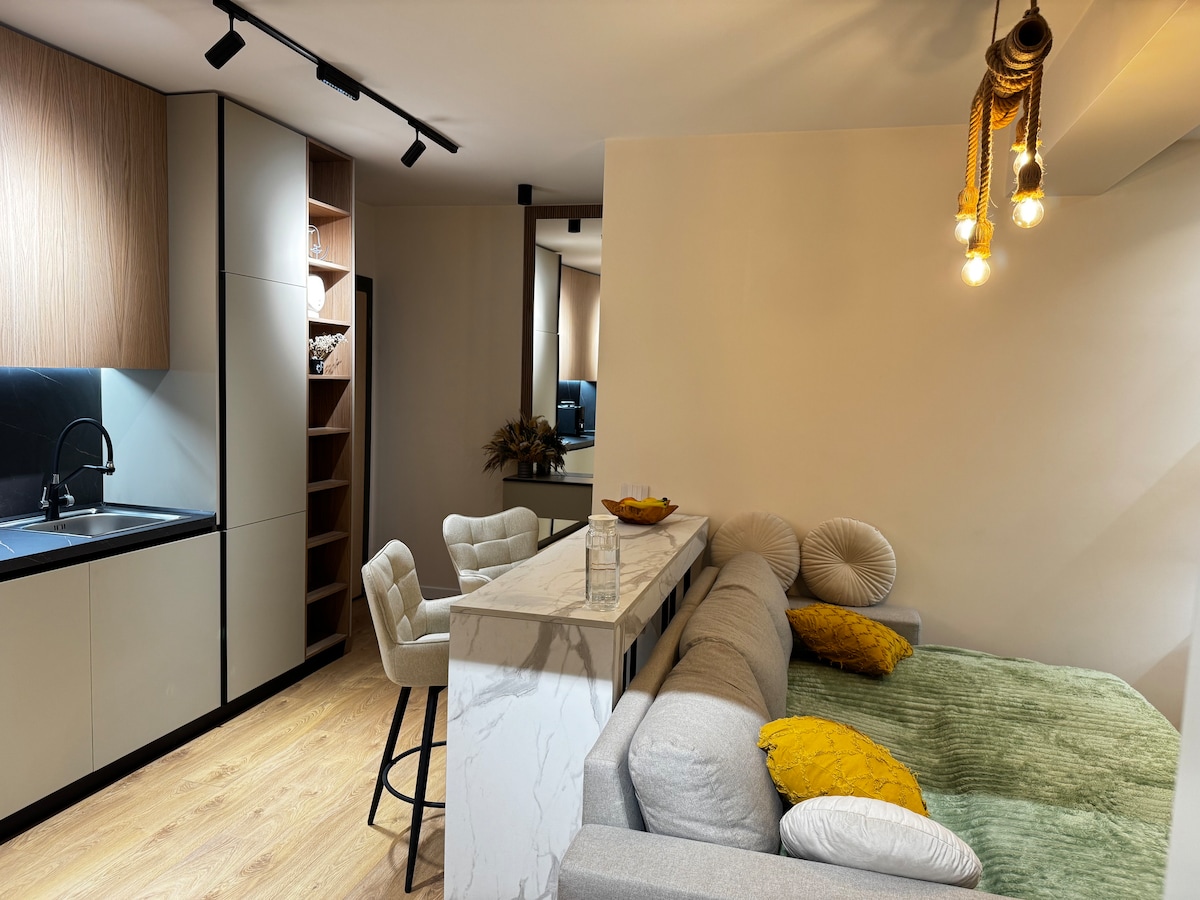 A modern kitchen area is seen, featuring wood and neutral-toned cabinetry. A sleek countertop provides space for dining, complemented by plush bar stools. A cozy living space is present, adorned with soft pillows and a vibrant green throw on a sectional sofa.
