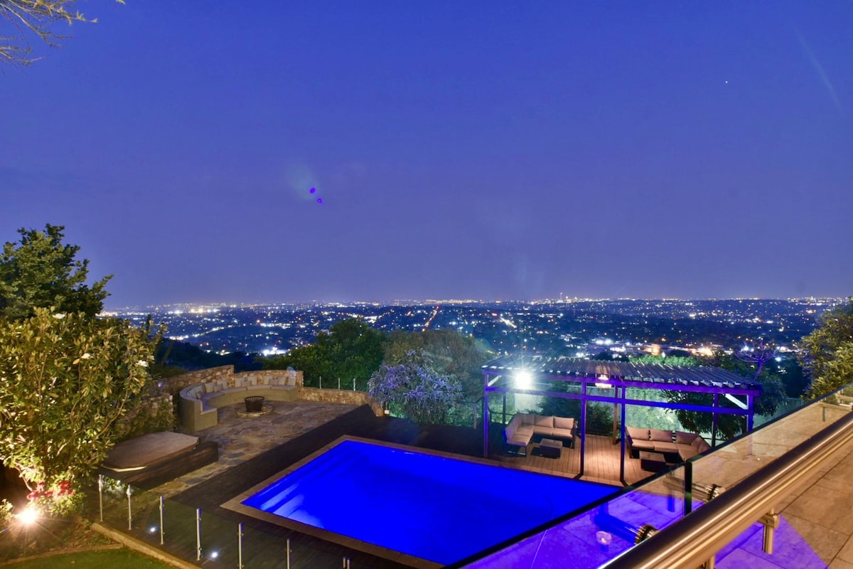 This evening view captures a shimmering blue pool complemented by seating under a pergola. The expansive city skyline of Johannesburg is visible in the background, illuminated against the night sky, offering a serene and tranquil atmosphere for outdoor relaxation.