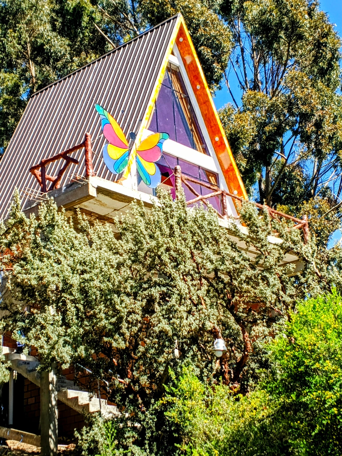 A distinctive A-frame house is set among lush greenery, featuring a large, colorful butterfly decoration on the front. The upper level includes a balcony with wooden railings, surrounded by trees, offering a sense of seclusion within a vibrant natural setting.