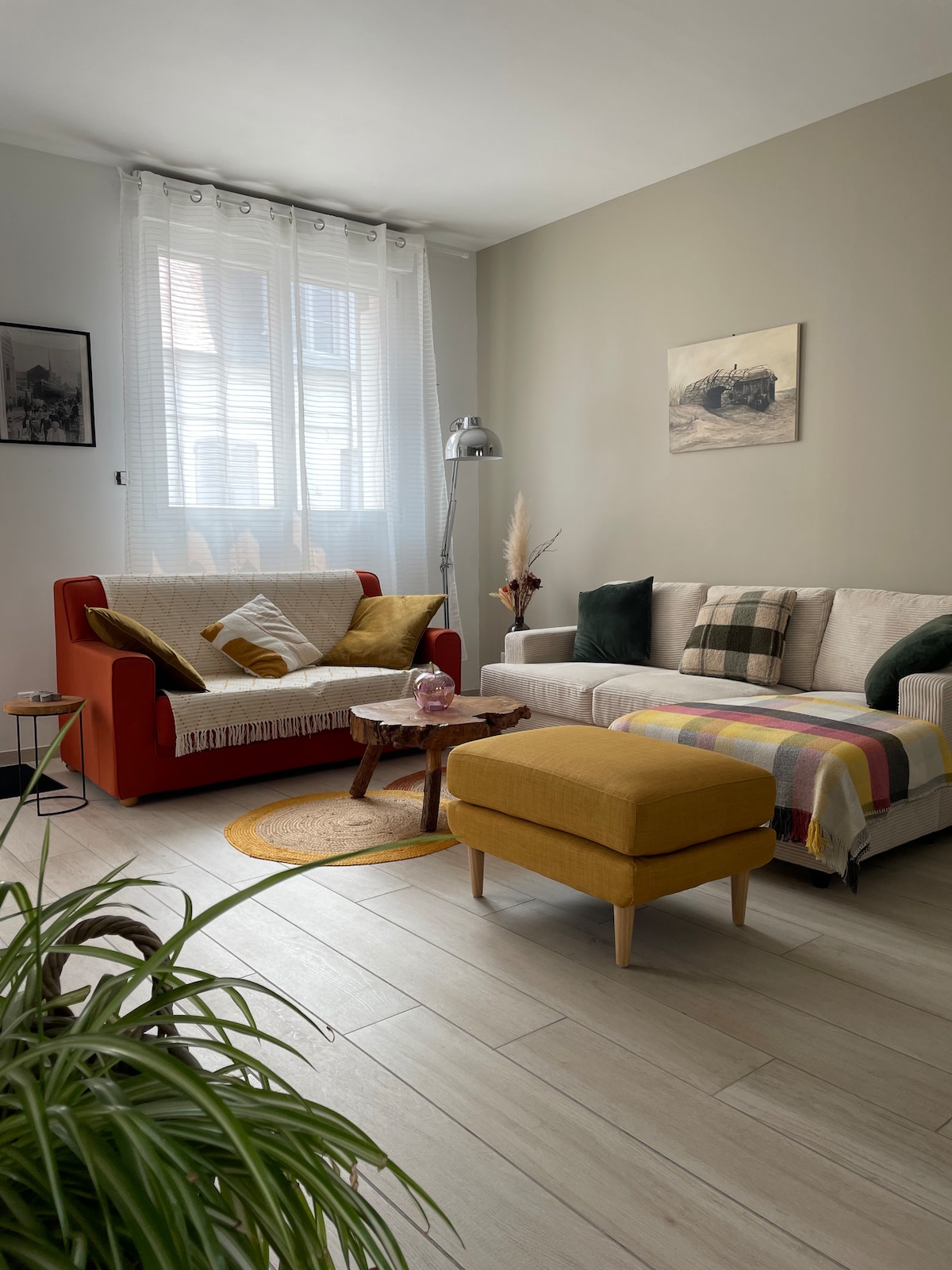 A cozy living area features two sofas, one in red and the other in light fabric, adorned with cushions. A round, woven rug sits under a small wooden table. Natural light filters through sheer curtains, while a floor lamp and decorative plants add warmth to the space.