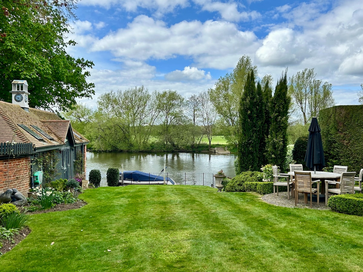 A lush green garden is seen beside a calm river, with neatly trimmed grass and leafy trees framing the view. A dining area is arranged with wooden chairs around a round table, and a large umbrella stands nearby, offering shade. Soft clouds drift across the blue sky.
