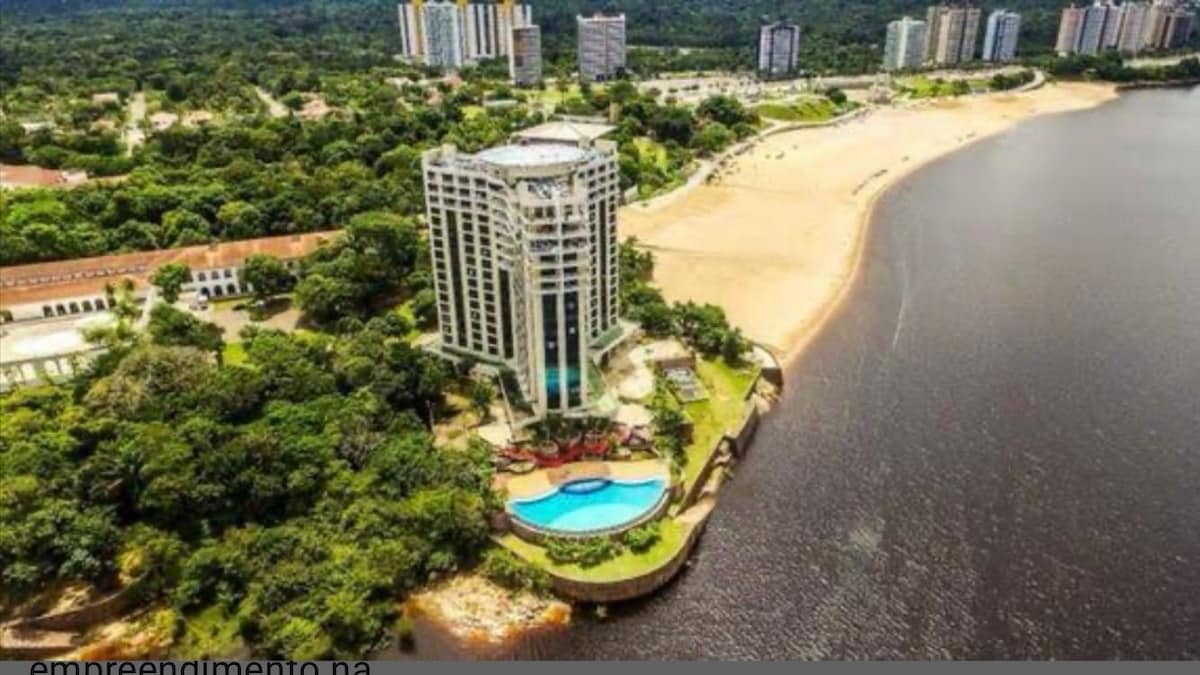 An aerial view captures a high-rise building surrounded by lush greenery and a sandy beach adjacent to a river. A swimming pool is visible at the base of the structure, with the contrasting colors of the water and landscape creating a serene environment.