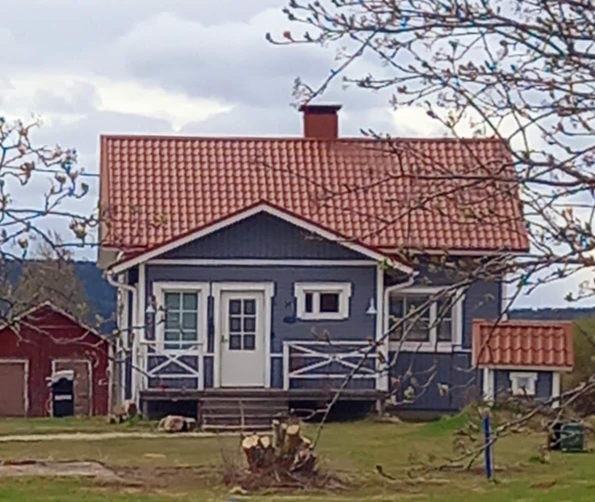 A two-story blue house features a red-tiled roof and white trim around the windows and doors. A front porch with railings offers access to the entrance. A small garden area includes logs and is surrounded by trees, adding to the rural charm of the setting.
