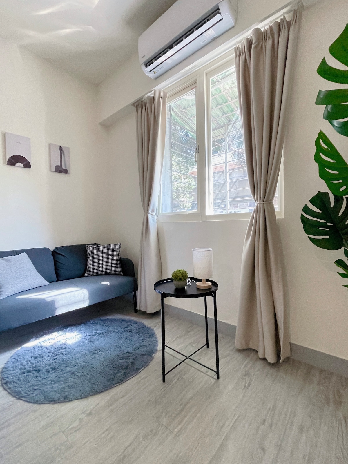 A cozy sitting area features a dark sofa adorned with light gray cushions. A small round table is positioned nearby, displaying a candle and a decorative plant. Natural light filters through the window, partially covered by beige curtains, enhancing the calming ambiance.