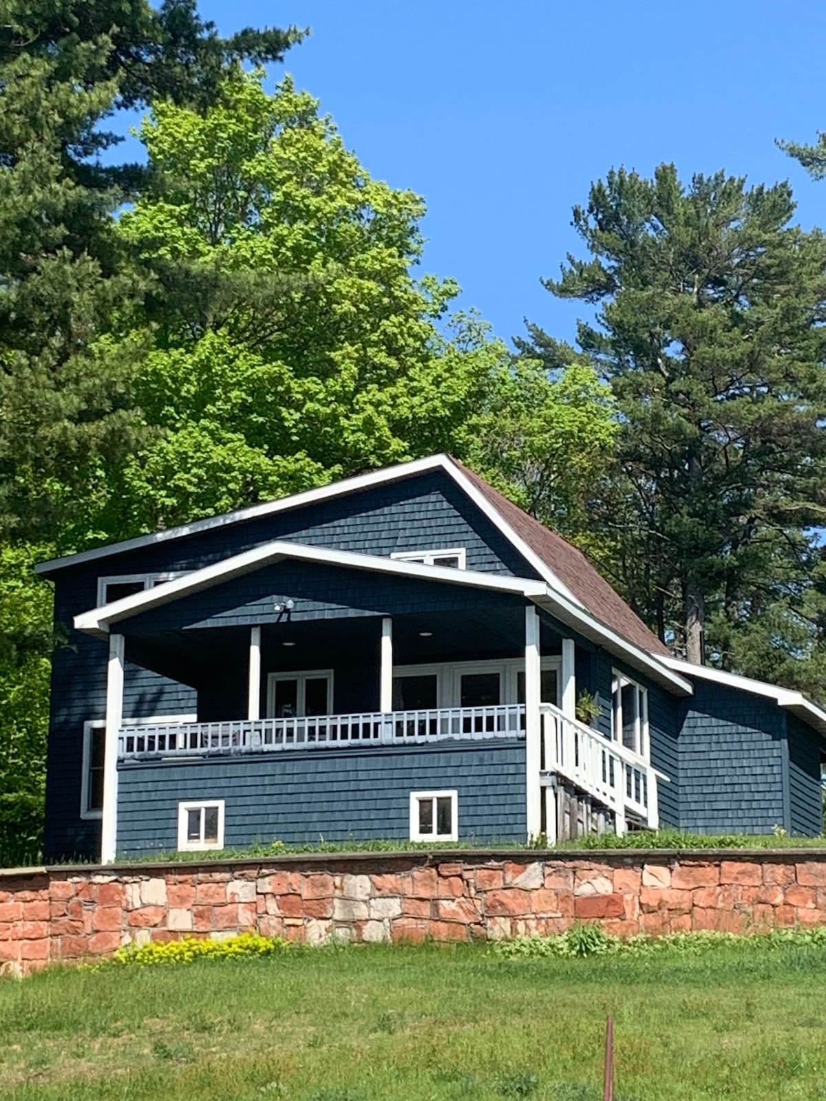 The home is depicted from a slight distance, showcasing its blue exterior and white trim. A wraparound porch is present, providing a place to relax. Lush green trees surround the property, enhancing its natural setting against a clear blue sky.