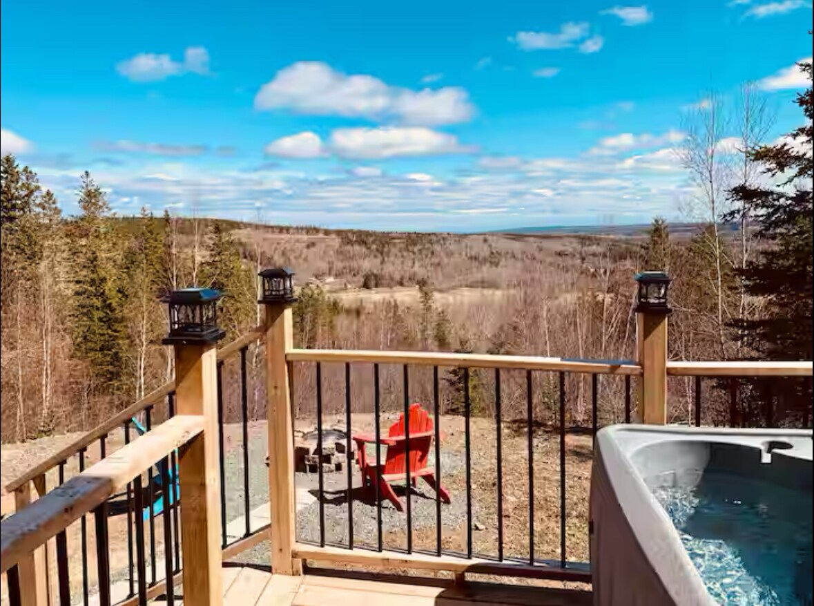 A scenic view from the deck offers expansive landscapes, featuring a fire pit surrounded by red adirondack chairs. The private hot tub is positioned near the railing, with trees in the background under a clear blue sky.