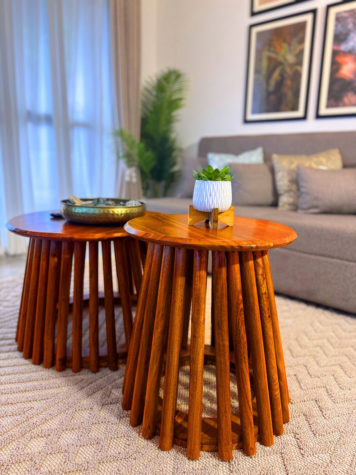 Two wooden side tables are positioned beside a modern sofa, featuring a circular top and uniquely designed legs. A small potted plant adds a touch of greenery to one table, while a decorative bowl rests on the other. Soft natural light filters through sheer curtains.