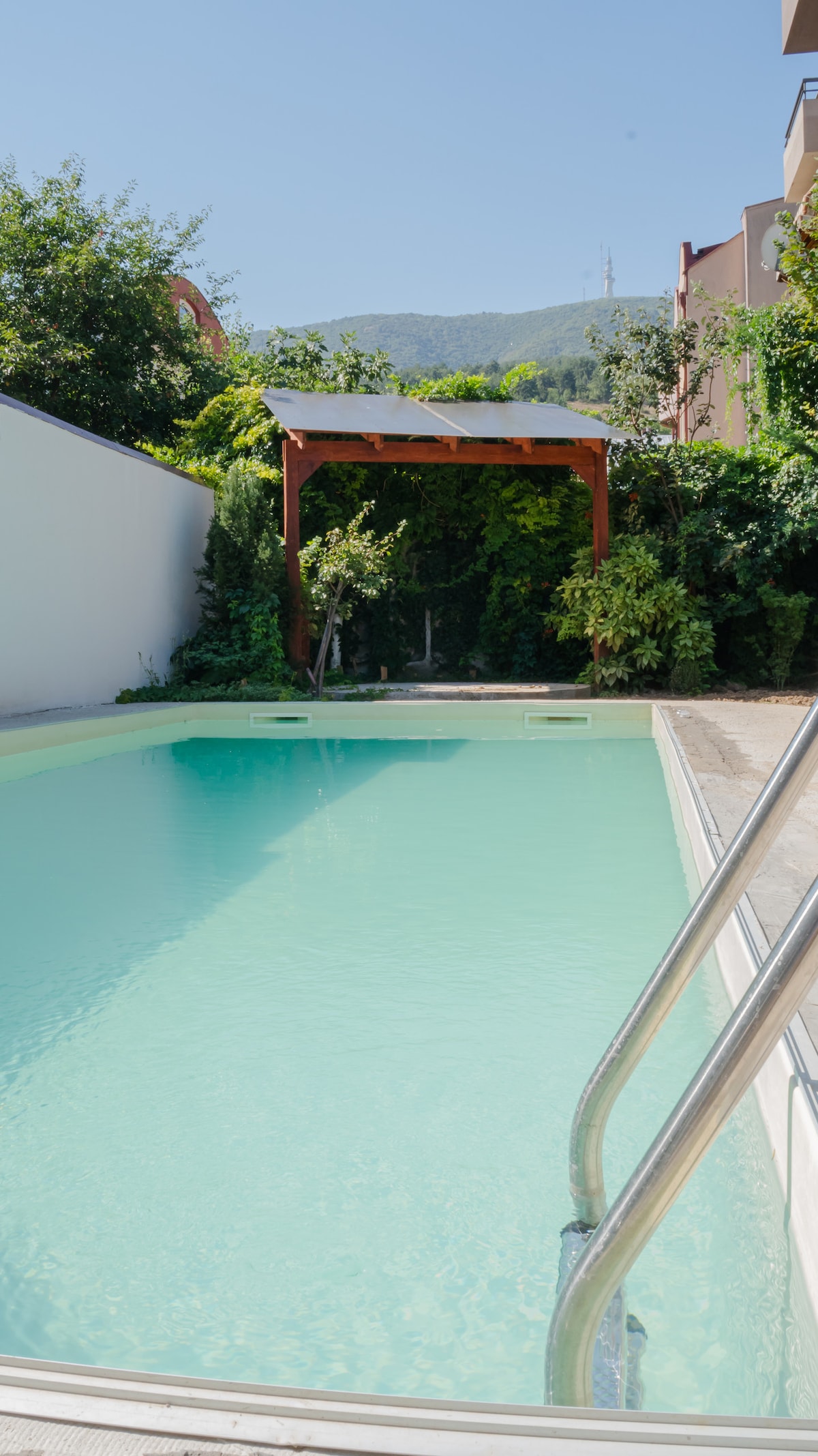 An outdoor pool features clear water and is surrounded by lush greenery. A wooden pergola provides shaded seating nearby, adding a natural touch to the tranquil setting. The pool area is designed for relaxation in a peaceful environment.