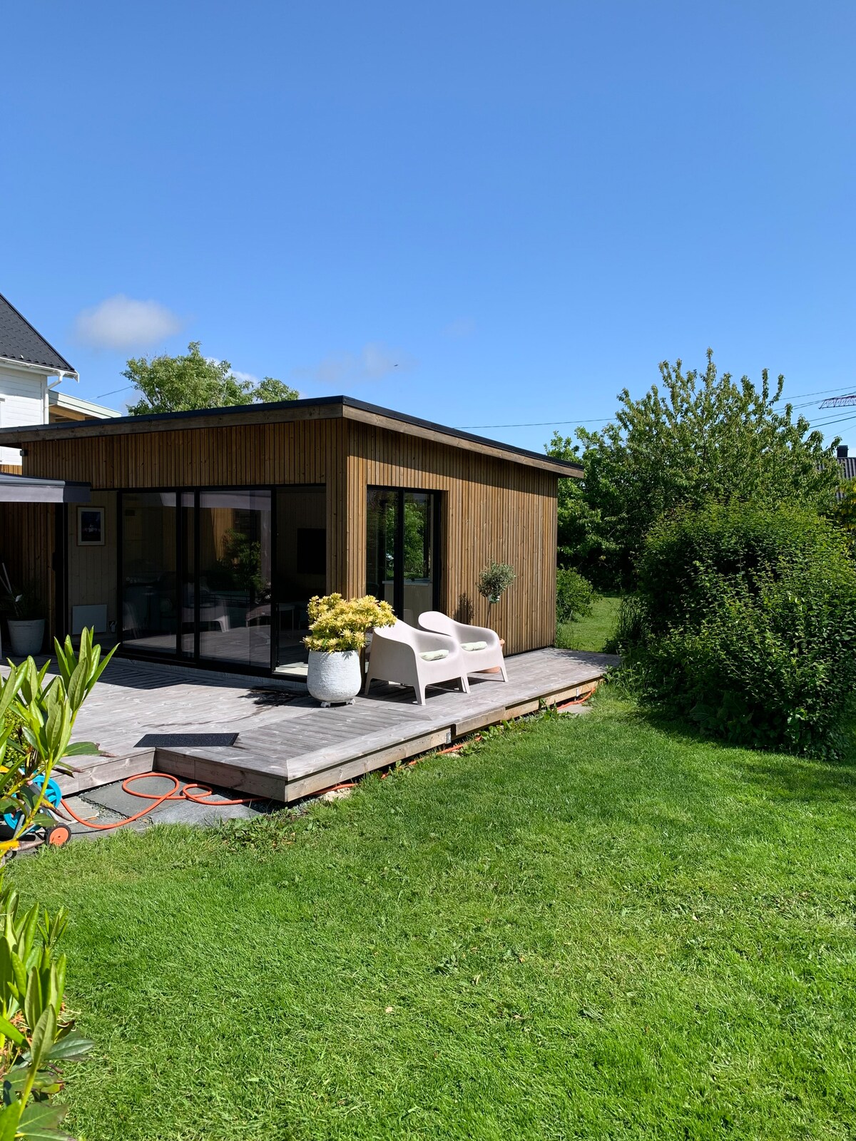 A wood-clad building features large glass doors that open to a spacious outdoor deck. A white chair sits beside a planter filled with yellow flowers. Lush green grass surrounds the area, and trees provide a natural backdrop under a clear blue sky.