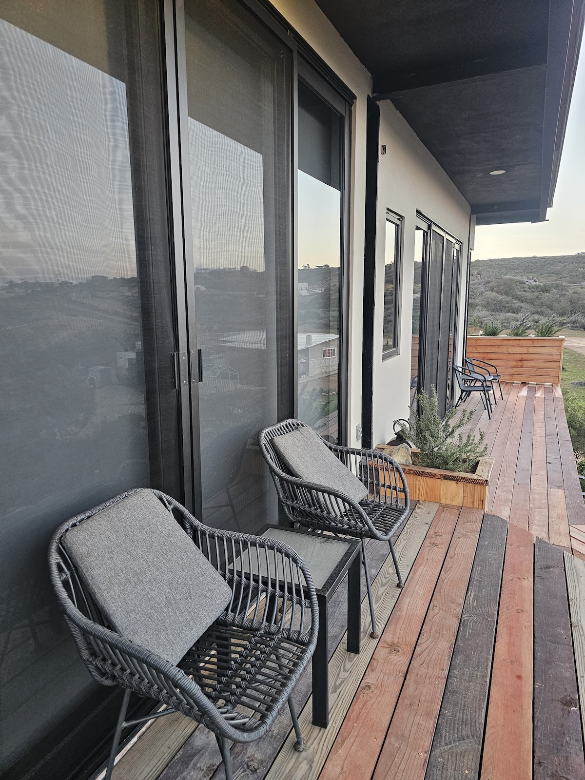 A wooden deck area features several black wicker chairs with gray cushions, arranged alongside sliding glass doors. A landscaped area with greenery is visible, complementing the serene environment.