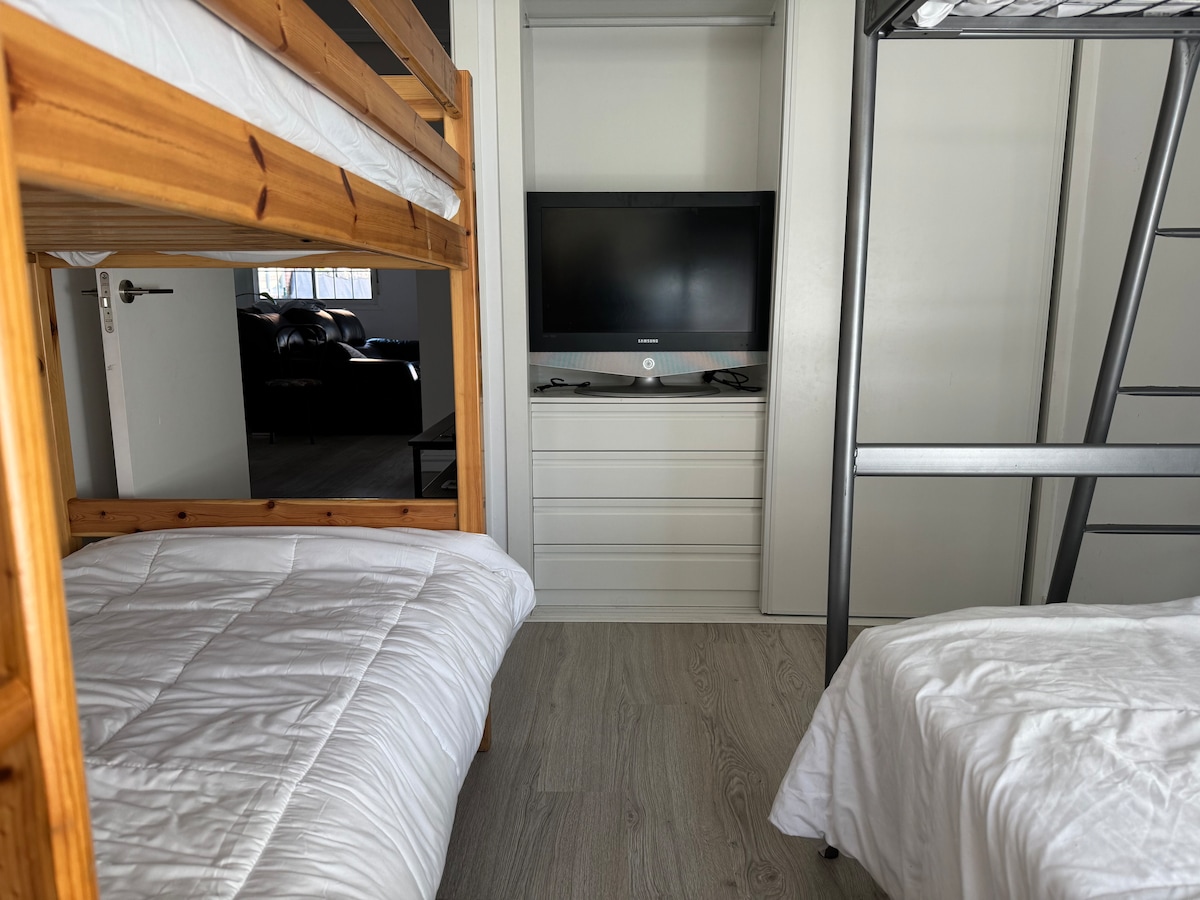 A cozy sleeping area features a wooden bunk bed alongside a separate single bed, each made with white linens. A wall-mounted television is positioned across from the beds, with a set of drawers visible in the background. Natural light enters through a nearby window.