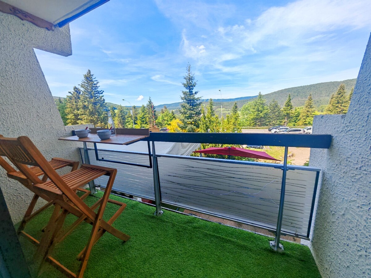 A balcony is positioned off the living area, featuring a wooden chair and a small table. Scenic views of trees and mountains are visible in the background, with a clear blue sky above. The space is carpeted with green outdoor grass.