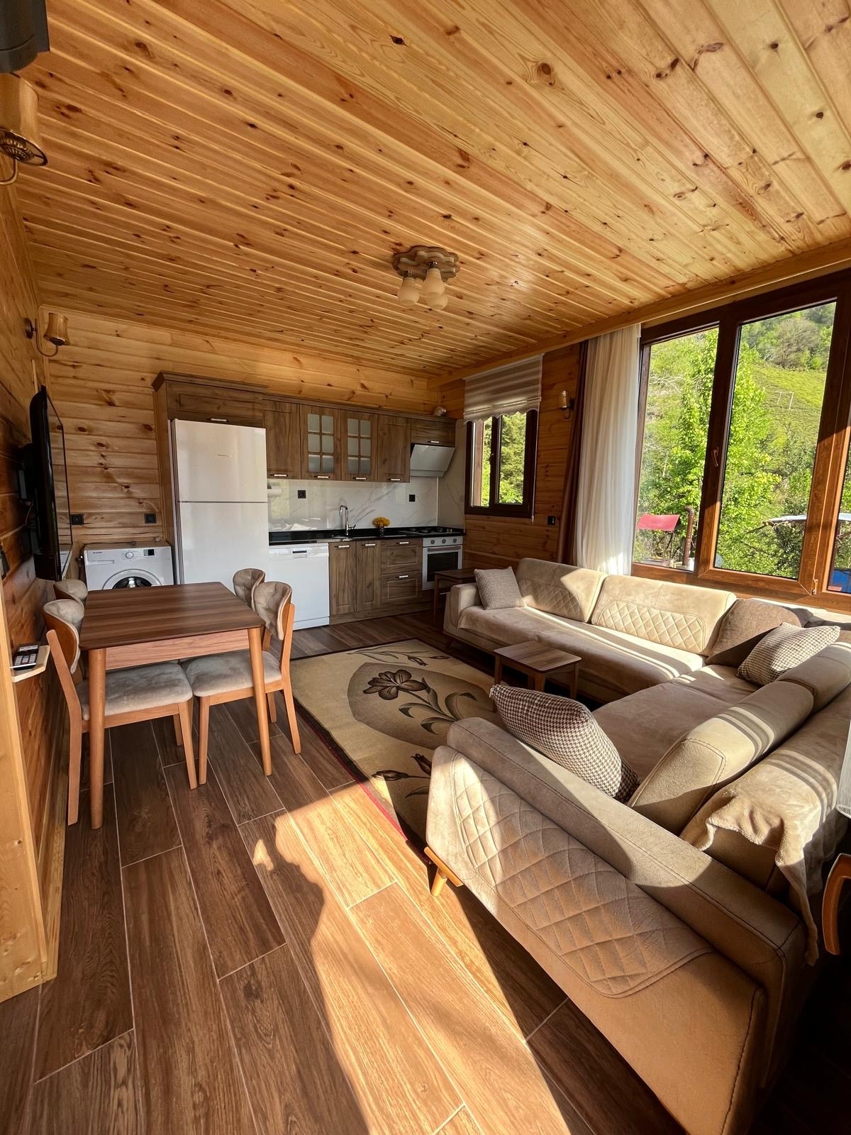 A warm interior is featured, showcasing wooden panel walls and a ceiling with exposed beams. A comfortable sofa and dining area are positioned near large windows that allow natural light to illuminate the space. A fully equipped kitchen is visible in the background.