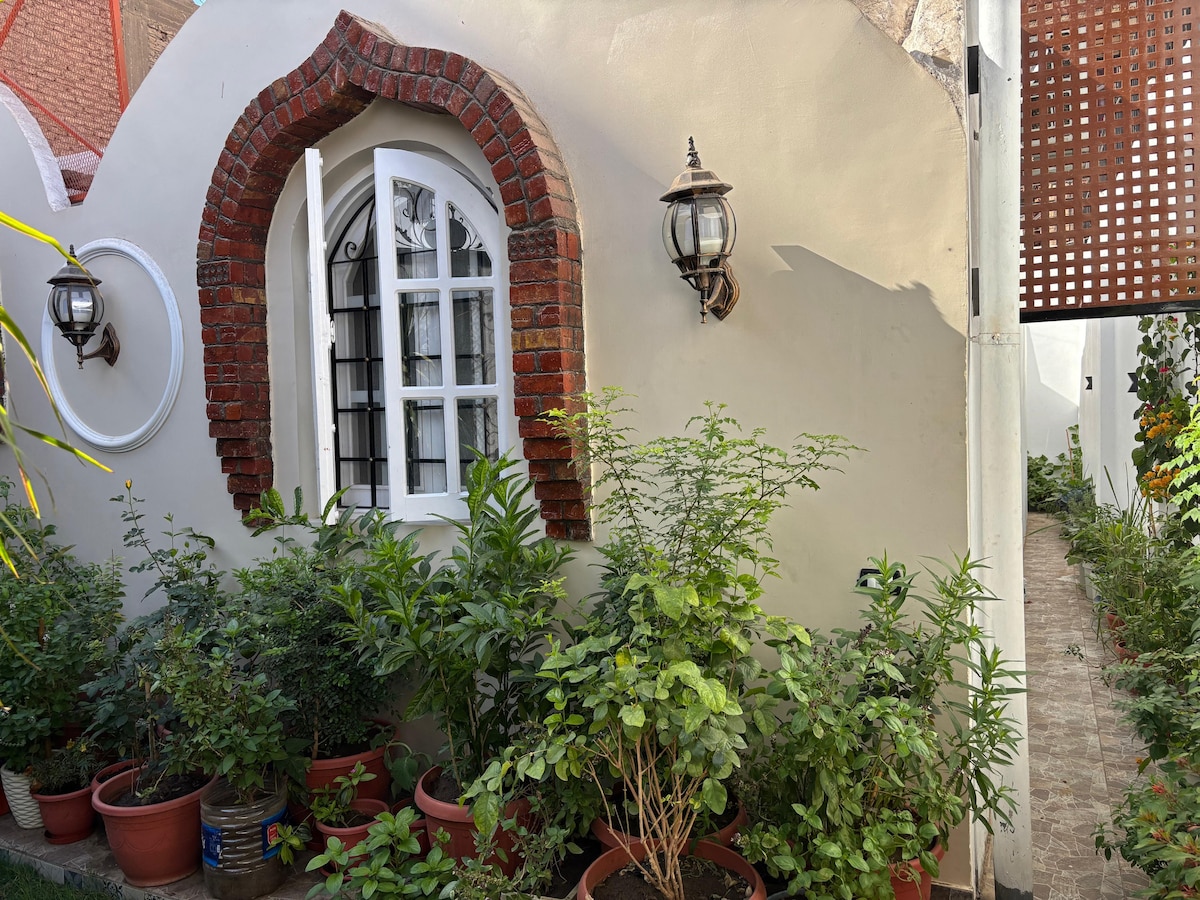 A charming exterior of the guest house is displayed, featuring a white wall with a decorative arched window framed by red brick. Lush greenery in potted plants surrounds the area, while a lantern adds a warm touch, enhancing the inviting atmosphere.