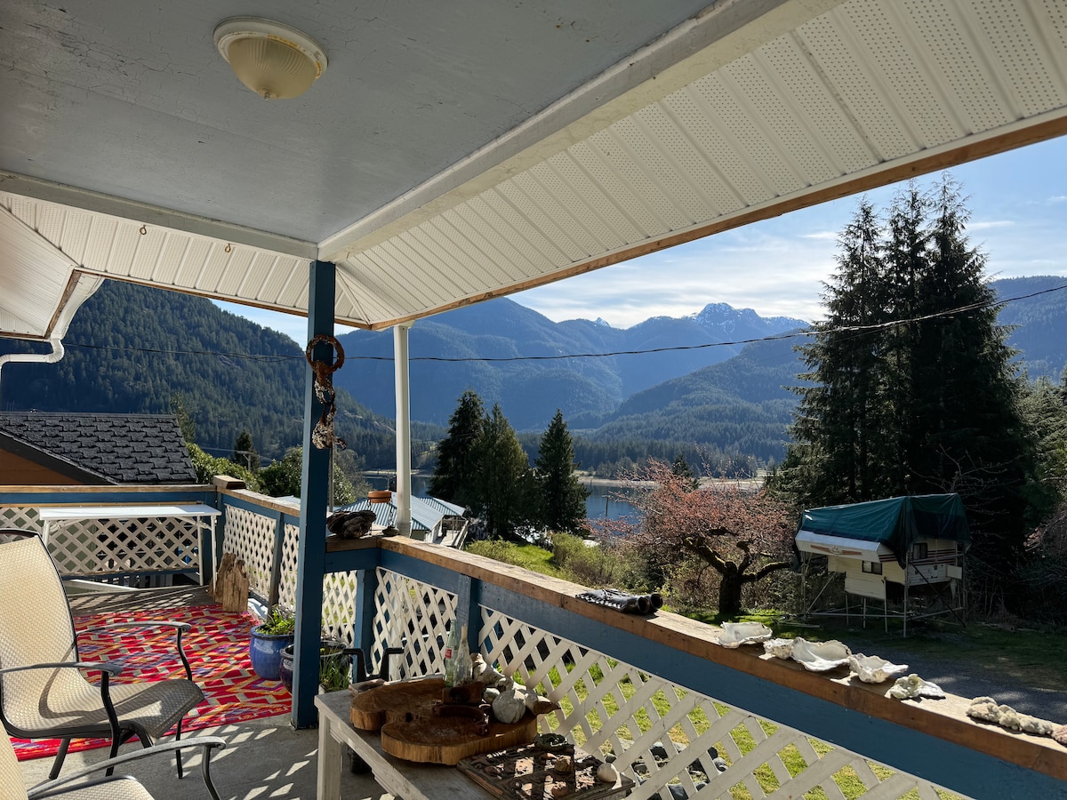 A covered porch is shown with two chairs facing out towards a scenic view of mountains and a serene inlet. The space is accessorized with various decorative items and a colorful rug, inviting relaxation while enjoying nature.