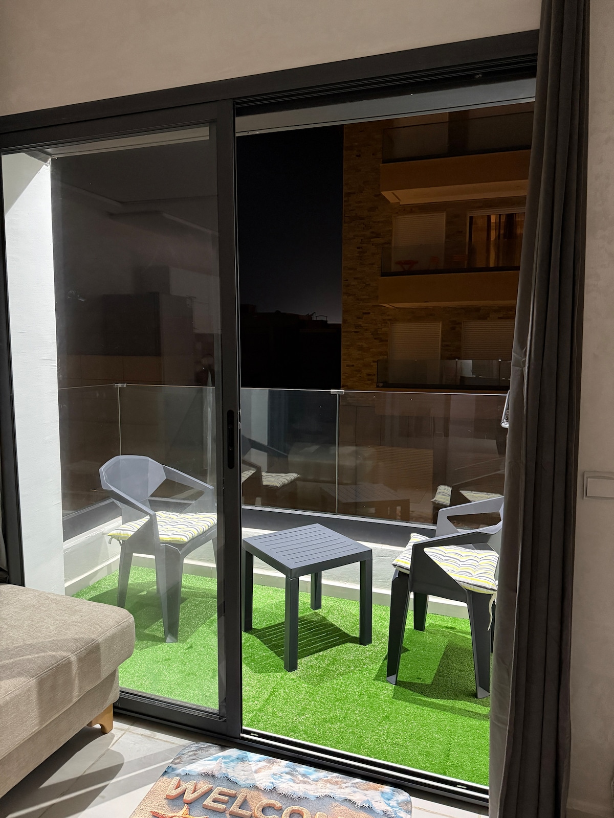 A balcony area is visible through sliding glass doors, featuring two chairs and a small table situated on artificial grass. Soft outdoor lighting highlights the space, creating a relaxed atmosphere for enjoying evening views.