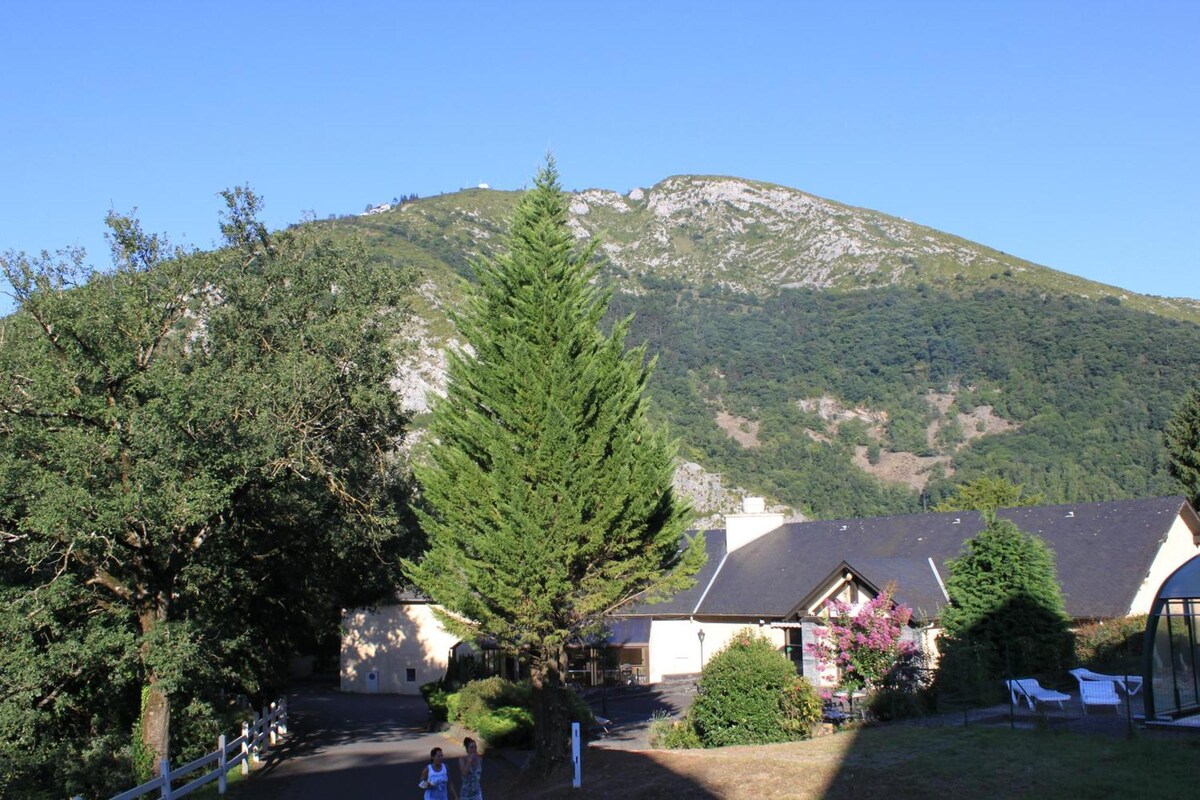 A serene landscape features a hillside, prominently displaying lush greenery and a nearby mountain peak. The area includes a modest residential building surrounded by trees and shrubs. A clear blue sky adds to the tranquil ambiance, suggesting a peaceful environment for relaxation.