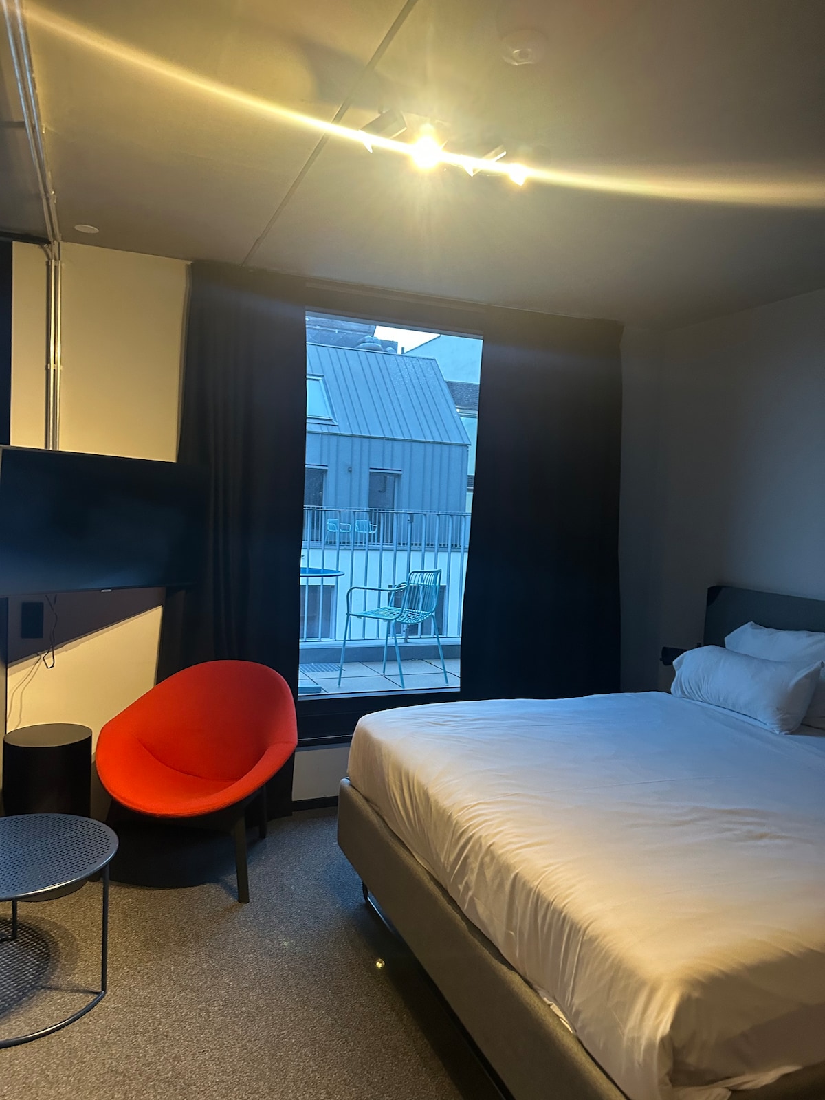 A modern studio room features a spacious king-size bed dressed in crisp white linens. A vibrant red chair adds a pop of color next to a small round table. A large window allows natural light to fill the room, with dark curtains framing the view.