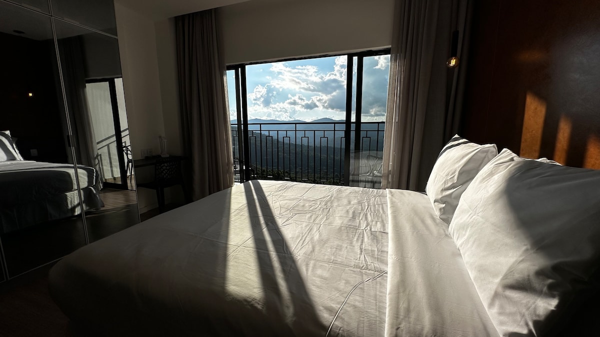 A well-lit bedroom features a spacious bed with crisp white linens. Large windows offer a scenic view of clouds and mountains in the distance. Soft natural light casts gentle shadows across the room, enhancing the serene environment.