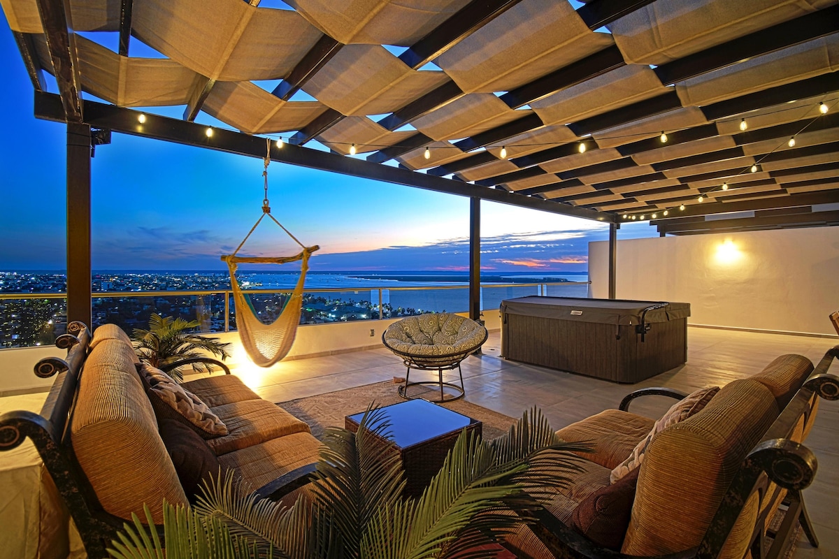 The private rooftop terrace showcases a comfortable lounging area with a large sofa, hammock, and lounge chairs. Low lighting beneath the pergola enhances the evening ambiance, while breathtaking views of the ocean and city skyline create a serene backdrop.