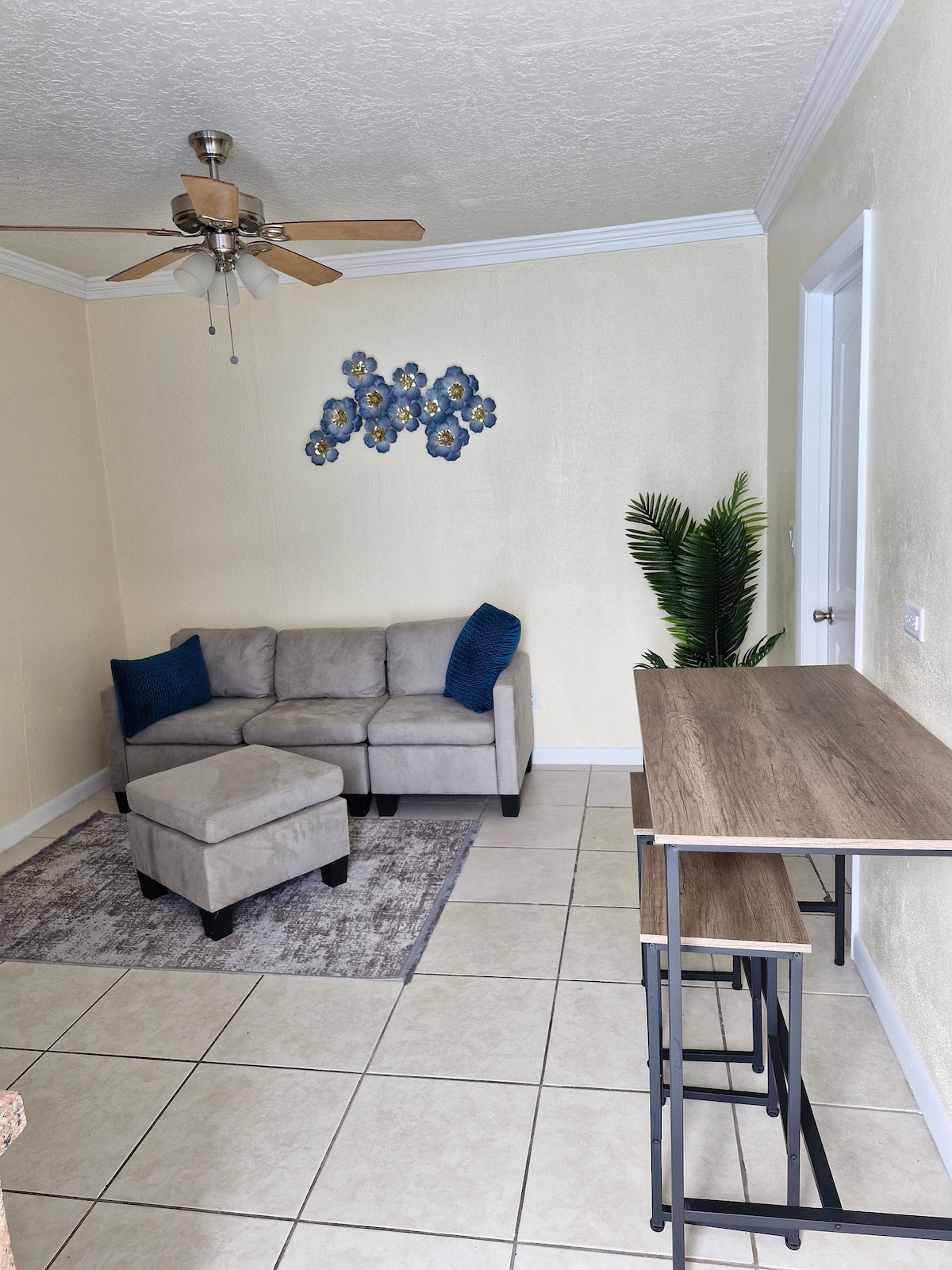 A cozy living area is presented, featuring a light gray sectional sofa with a matching ottoman. A small wooden table with high stools is positioned nearby. Decorative blue wall art is displayed above the seating, and a tall green plant adds a touch of nature to the space.