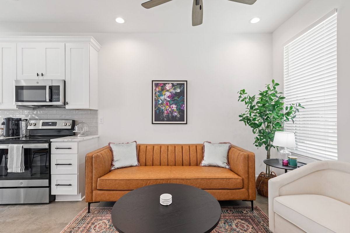 A modern living area features a rust-colored sofa with light cushions, a round black coffee table, and a soft area rug. Natural light enters through the window beside a small tree and a lamp, while a framed floral print adds color to the wall.