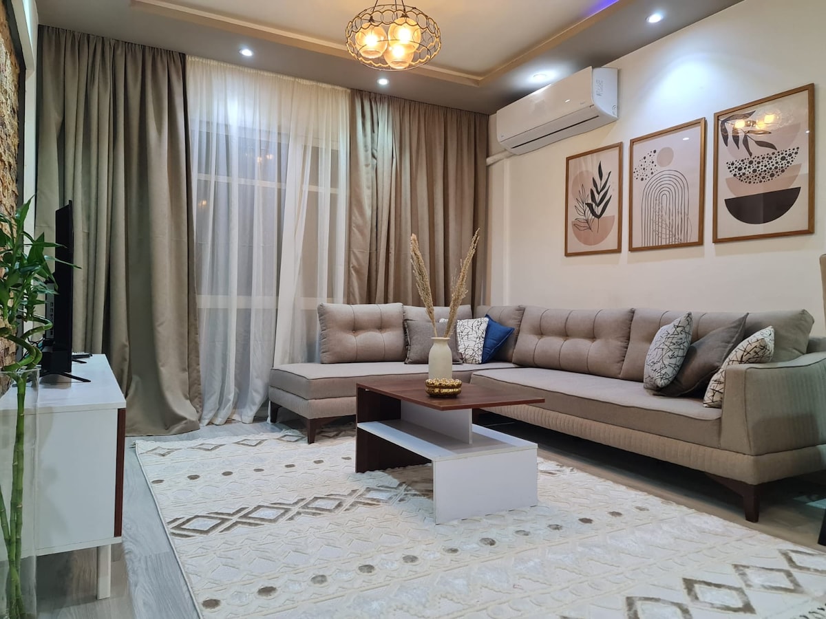 A modern living room features a sectional sofa adorned with decorative pillows, placed next to a sleek coffee table. Wall art pieces complement the soft color palette. Large curtains frame the window, allowing natural light to enhance the décor, while a plush area rug anchors the space.