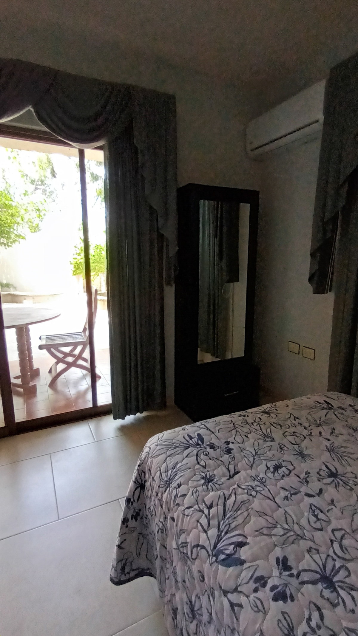 A bright bedroom area is equipped with a spacious bed adorned with a floral-patterned cover. A tall mirror cabinet stands nearby, reflecting the natural light from the glass doors that lead to a private outdoor seating area. Curtains frame the entrance, enhancing privacy.