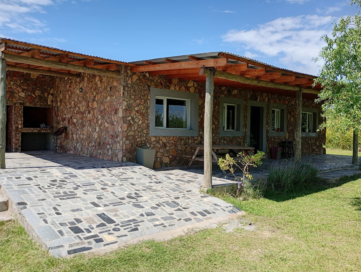 An exterior view of a spacious stone house is presented, featuring a covered porch supported by wooden beams. Smooth flagstone pathways lead to the entrance, with a grassy area and trees in the foreground, suggesting a serene outdoor setting.