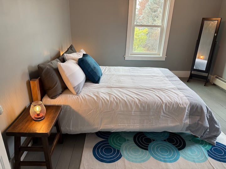 Chumba hiki cha kulala cha watu wazima kina kitanda cha ukubwa wa kifalme, mito ya kitani na mwonekano wa malisho ya amani nje ya dirisha. Kabati la nguo lililotengenezwa kwa mikono (halijaonyeshwa kwenye picha) linaongeza mvuto wa kijijini kwenye mapumziko haya tulivu, yaliyojaa mwanga.