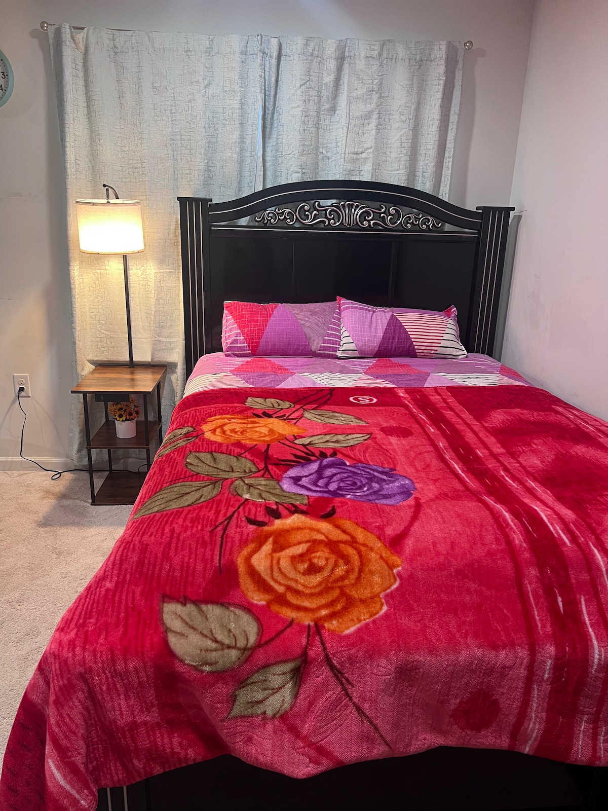 A spacious bed is dressed in a vibrant floral blanket, featuring roses in warm colors. Two decorative pillows rest against a patterned headboard. A nightstand with a lamp is positioned beside the bed, and light filters in through soft, light-colored curtains.
