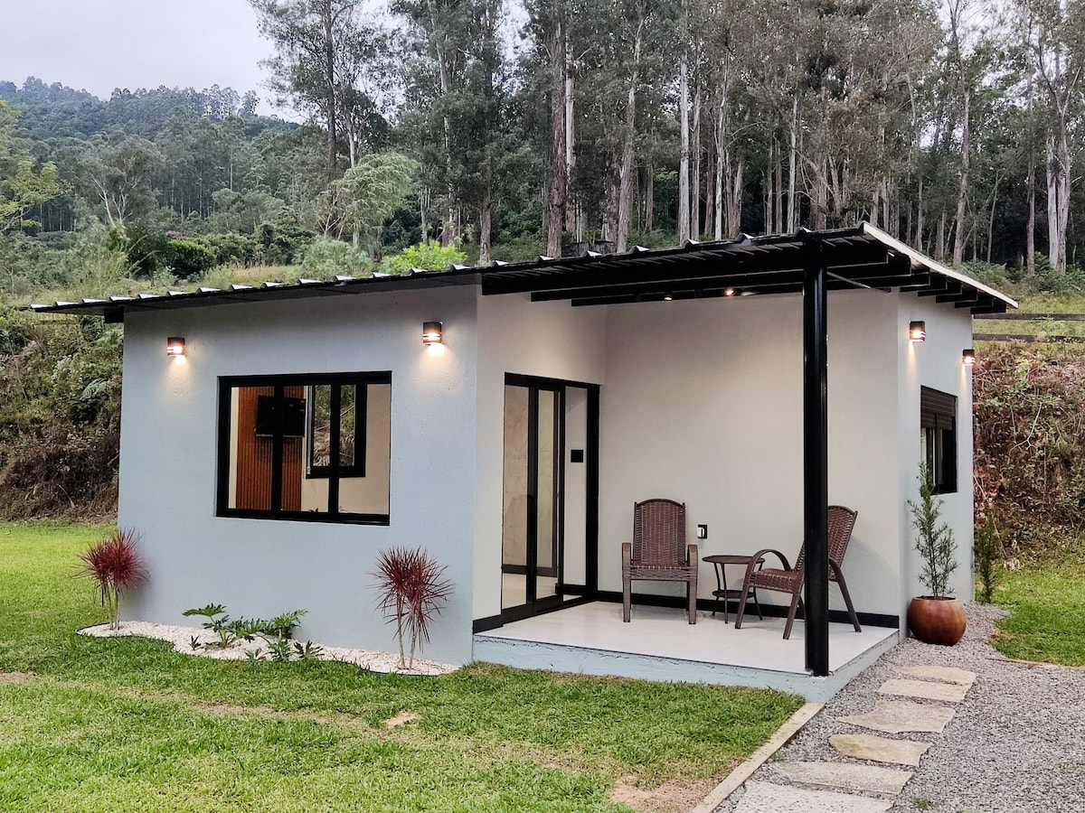 The exterior of the house features a simple, modern design with a light-colored facade. Two chairs are positioned under a covered porch, providing a welcoming entrance. Lush green grass surrounds the property, complemented by tall trees in the background, creating a natural, serene setting.