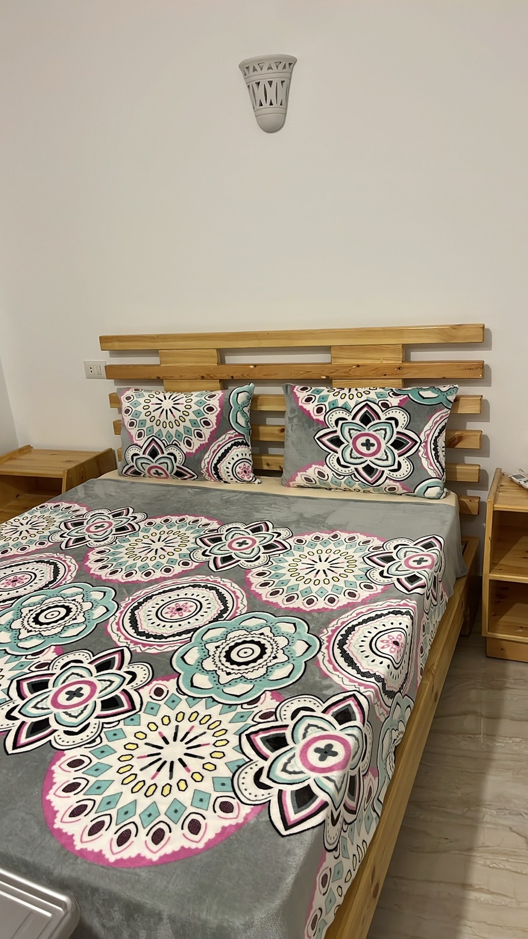 A wooden bed frame is depicted, featuring a colorful patterned bedspread with floral designs. Two matching pillows rest against the headboard. Nightstands are positioned on either side, contributing to a minimalist decor style while providing practical storage.