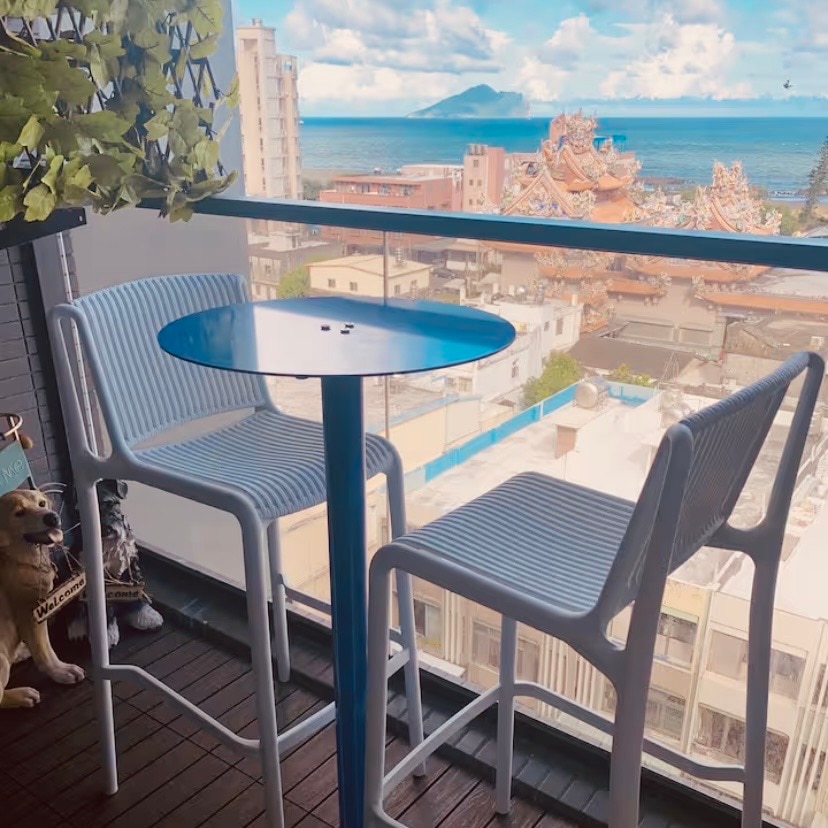 A small balcony space is furnished with a round blue table and two white bar stools. The view beyond features a vibrant coastal scene with rooftops and a distant mountain, while decorative plants hang from the railing, adding a touch of greenery.