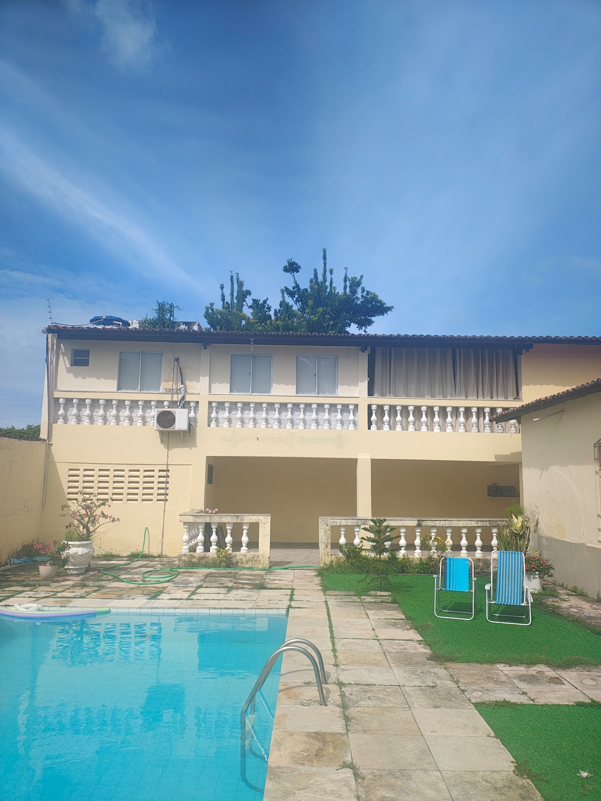 A two-story structure with a balcony overlooks a clean swimming pool. The patio features stone tiles and landscaped areas with greenery. Two lounge chairs are positioned near the water, while an air conditioning unit is visible on the wall, ensuring comfort in the warm surroundings.