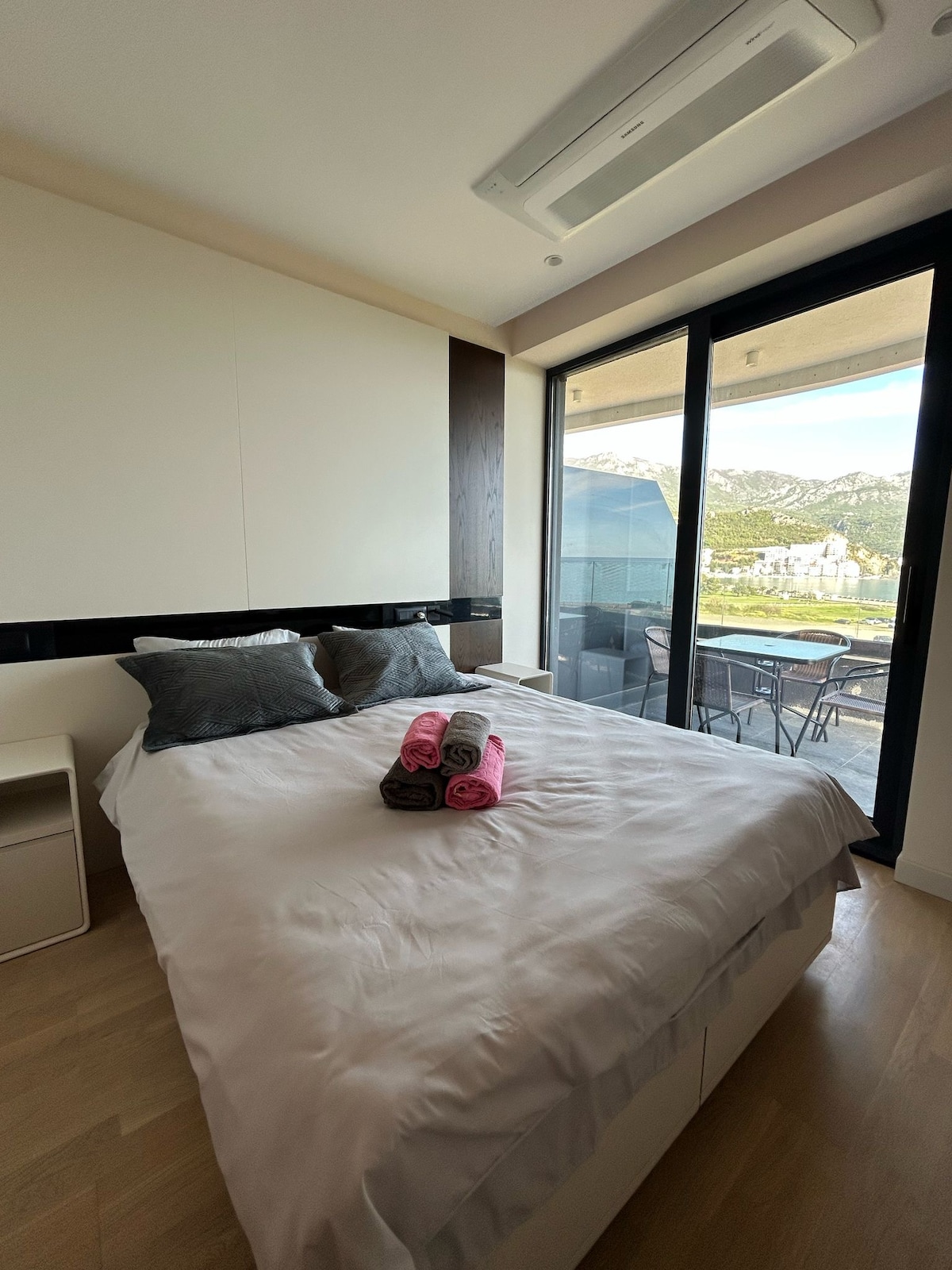 A well-appointed bedroom features a spacious bed topped with decorative pillows and rolled towels in shades of pink. Floor-to-ceiling windows provide access to a private balcony, offering views of the surrounding landscape.