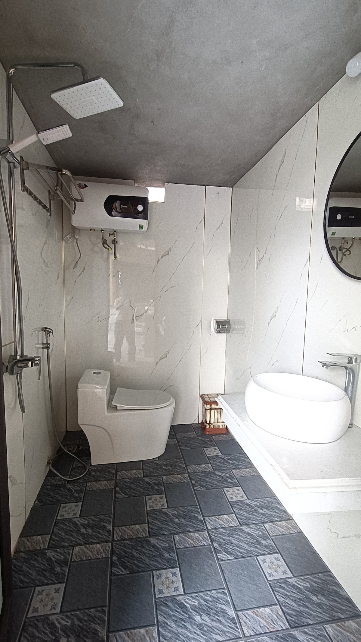 A modern bathroom features a white toilet and a basin on a stone countertop. A shower area with a rainfall showerhead is present, along with a round mirror reflecting ambient light. The floor is tiled with a dark patterned design, ensuring a clean and functional space.