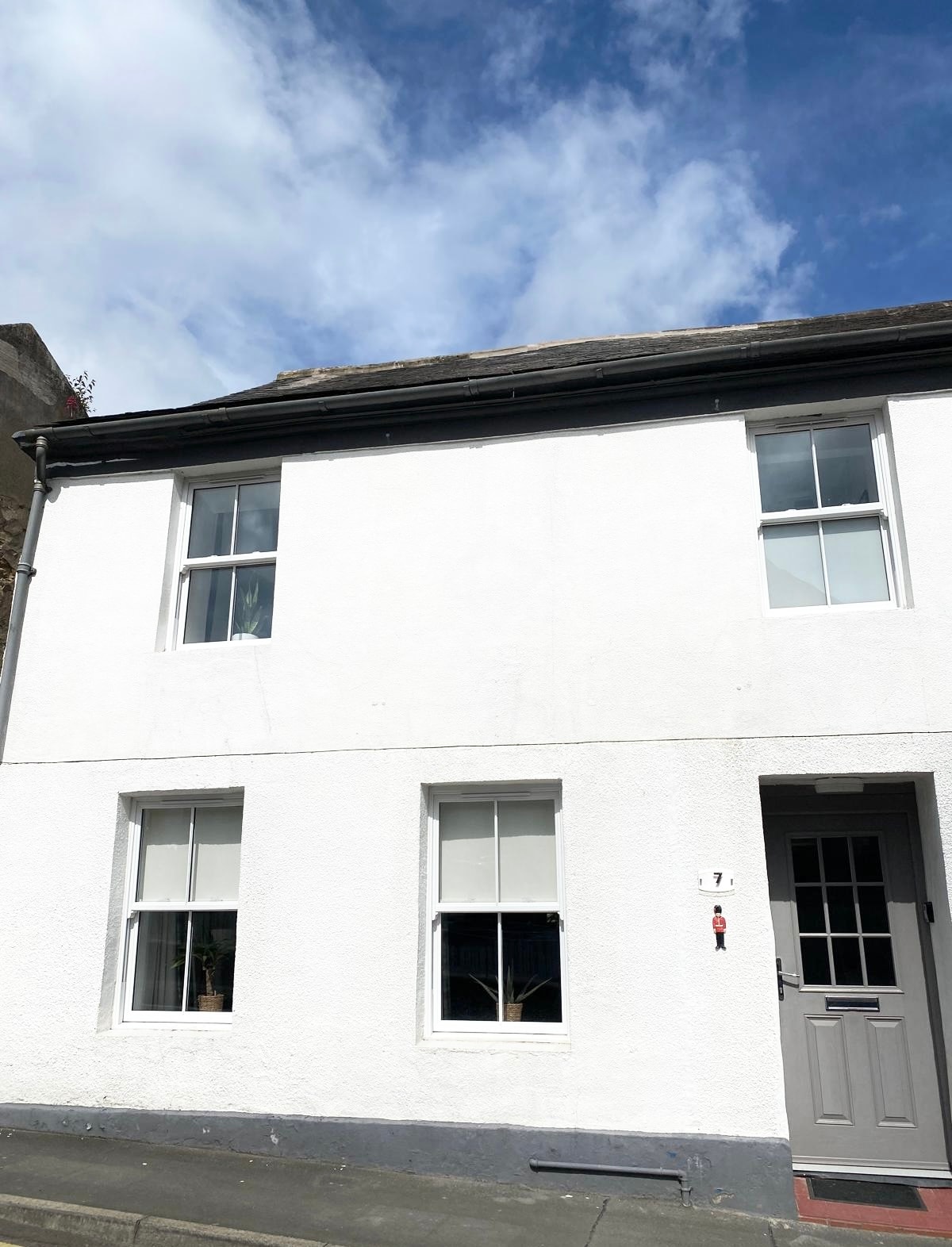 The exterior of a two-story white townhouse is displayed, featuring two symmetrical windows on the upper and lower levels. A welcoming front door is positioned to the right, complemented by a simple door knocker, under a partly cloudy sky.
