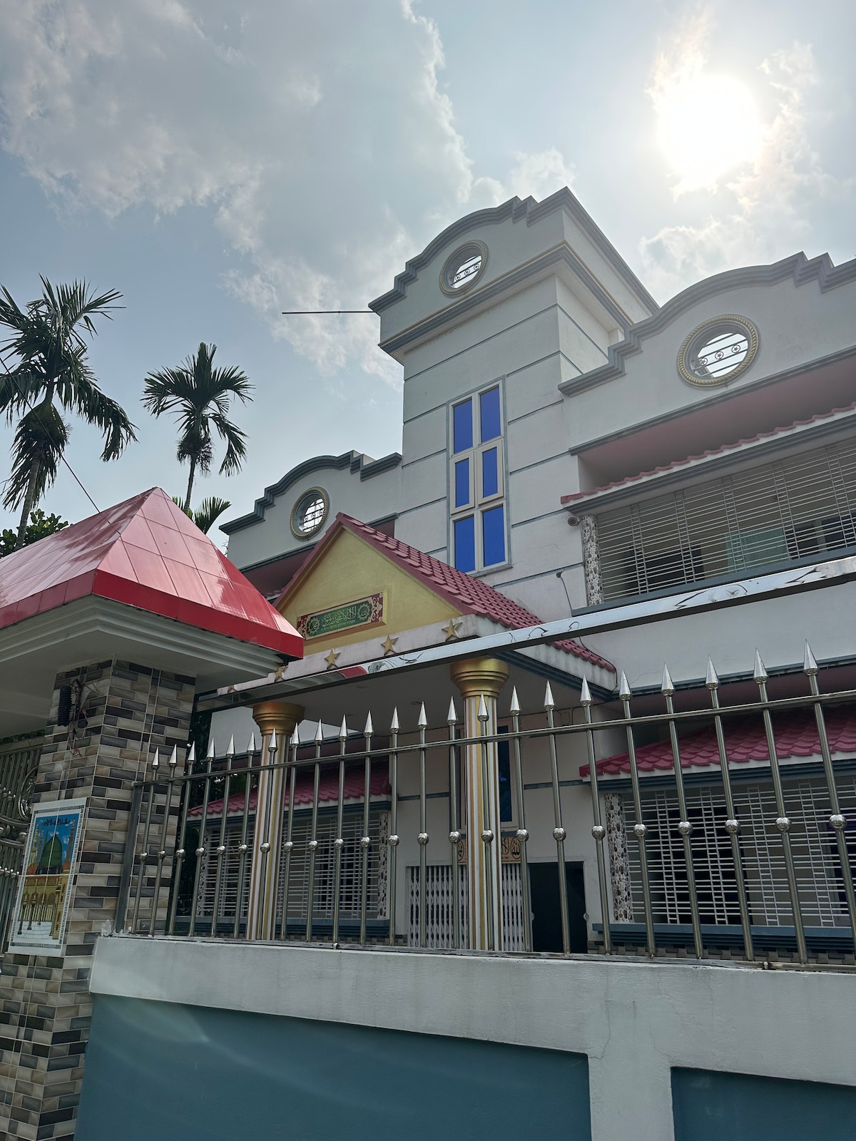 A three-story house is showcased with a distinct architectural design, featuring a red roof and blue accents. Tall palm trees frame the property, and a secure gated entrance adds to the privacy. Expansive windows allow natural light to enhance the exterior.