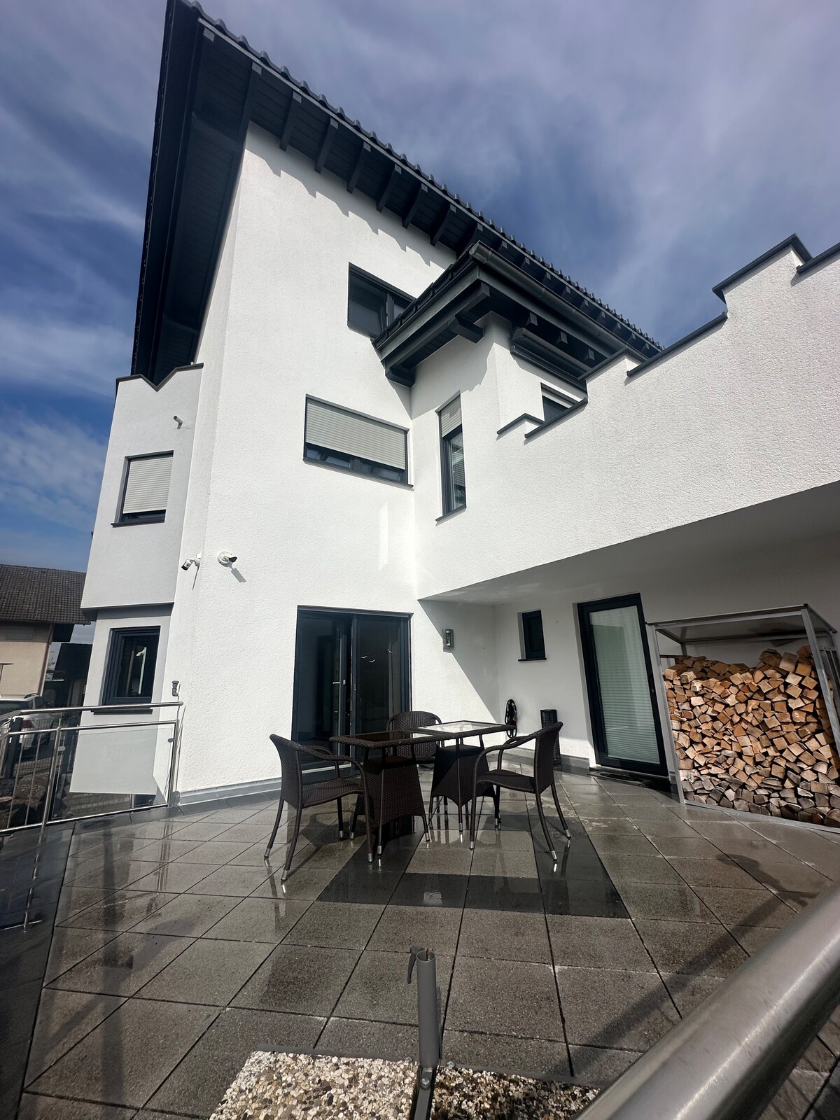 An outdoor patio is presented with a table and four chairs, set against a backdrop of a white building. A stack of firewood is neatly arranged nearby. The patio features sleek, polished tiles that reflect the surrounding natural light.