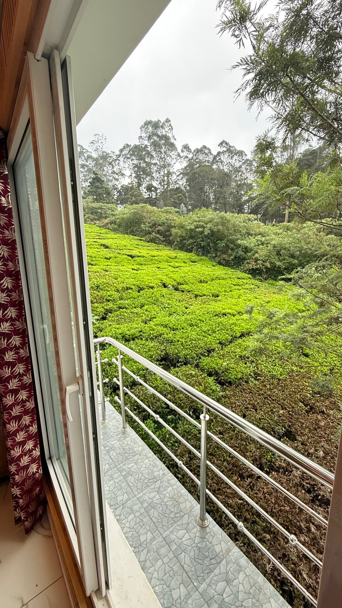 A balcony view overlooks lush green hills covered with tea plantations. The glass sliding door provides access to the outdoor space, while a patterned curtain partially frames the view. The texture of the plants is visible against a cloudy sky.