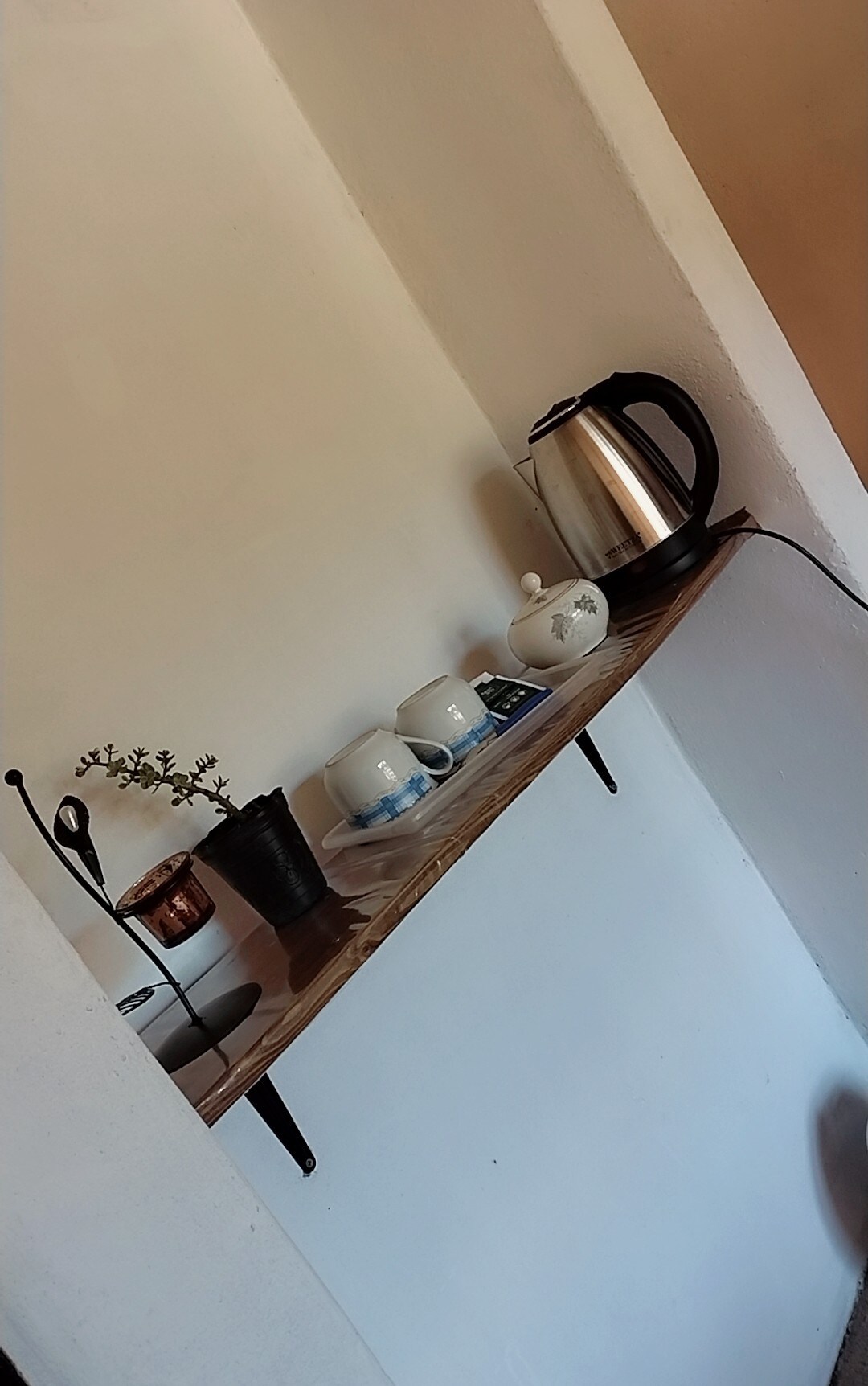 A simple shelf is mounted on a white wall, holding a kettle, teapot, and matching cups. A small vase with a plant adds a touch of greenery. Plates are neatly arranged alongside the items, creating a functional and inviting tea station.