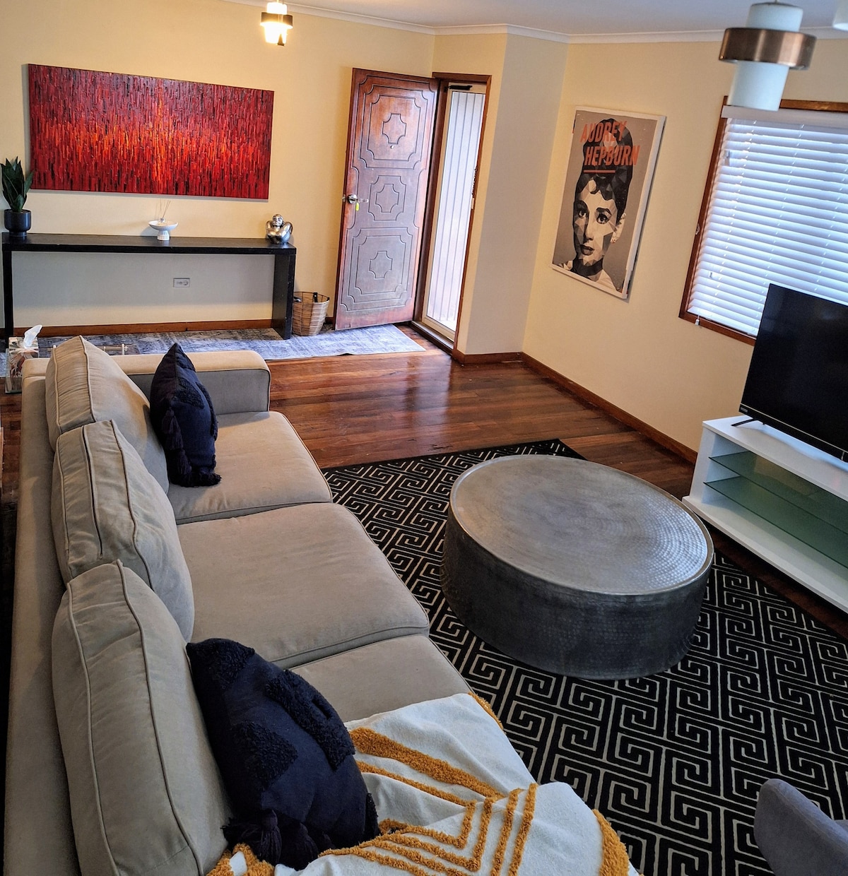 A comfortable living area features a spacious beige couch adorned with dark cushions and a patterned throw. A round coffee table sits atop a geometric rug, while natural light filters through the partially closed window. A decorative piece and two wall art prints enhance the atmosphere.