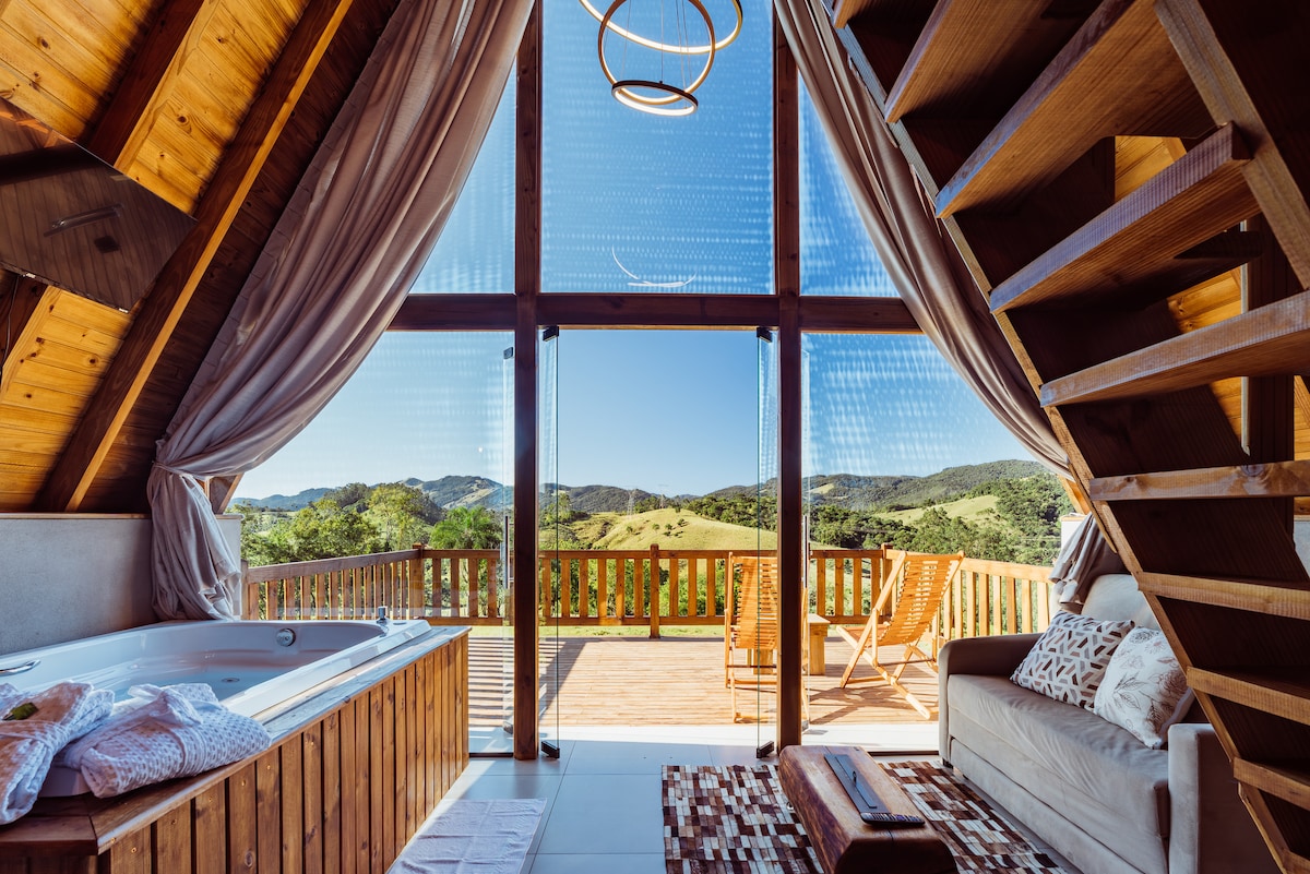 A spacious interior invites relaxation, featuring a cozy seating area and a spa tub near large glass windows. Natural light floods the space, revealing views of rolling hills and outdoor seating on a deck. The wooden staircase adds a rustic charm to the setting.