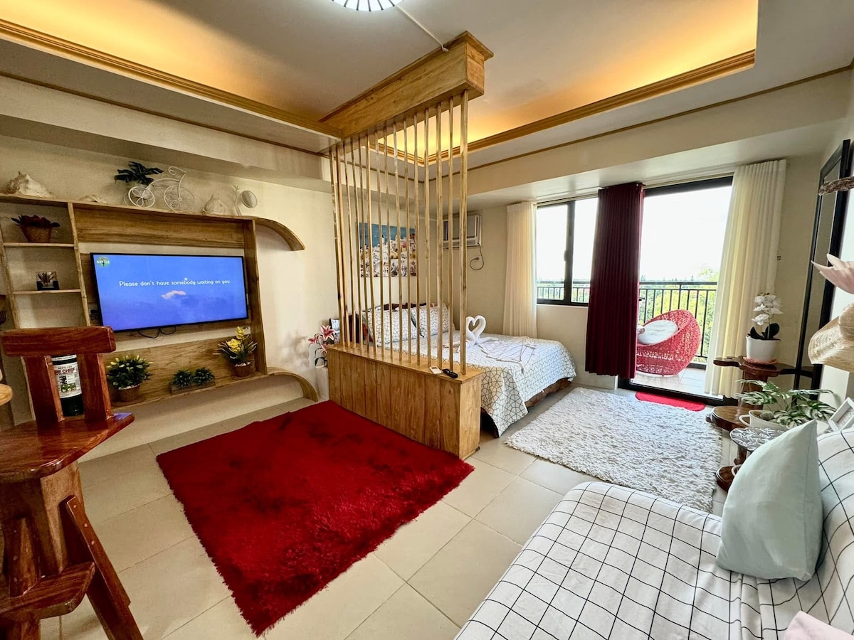 The interior showcases a split layout, featuring a cozy sitting area with a white couch and wooden accent table. A spacious bed is positioned behind a wooden screen, while a red rug adds warmth. Floor-to-ceiling windows open to a balcony view.