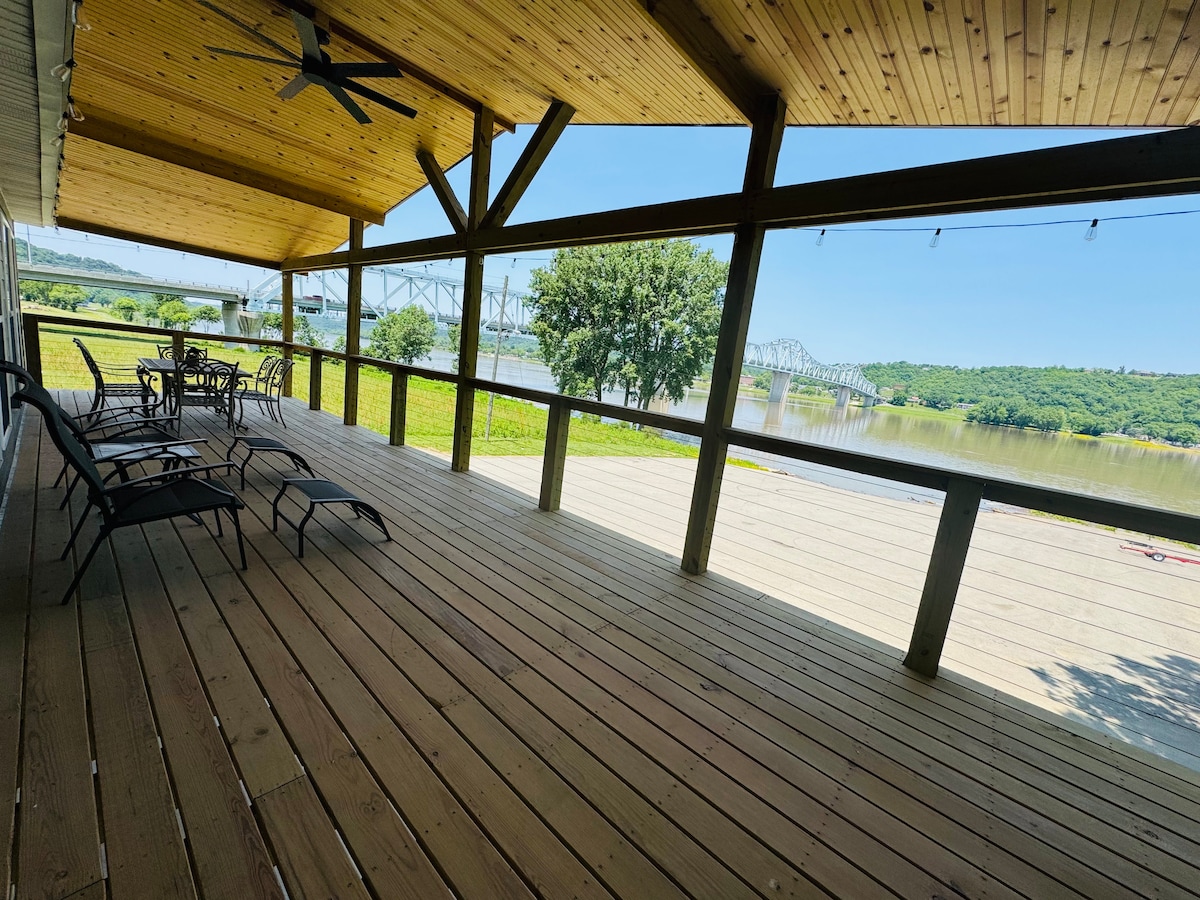 A spacious covered porch features wooden flooring and sturdy beams, providing an unobstructed view of the river and surrounding landscape. A table and several chairs are arranged for seating, while a ceiling fan is installed overhead for airflow.