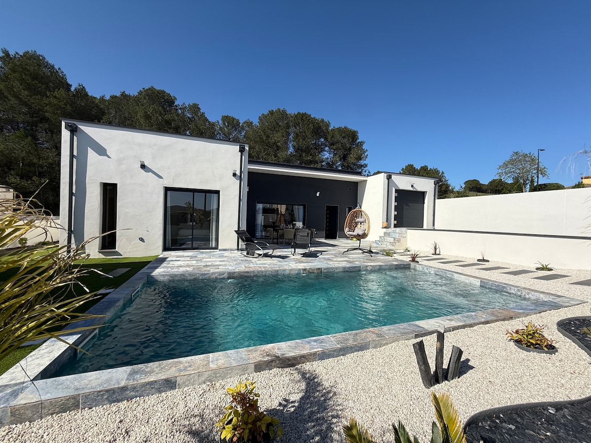 A modern villa is showcased with an inviting pool surrounded by a stone patio. The landscaped area features small plants and artificial grass. Large glass doors provide a view into the living space, enhancing the connection with the outdoor environment under a clear blue sky.