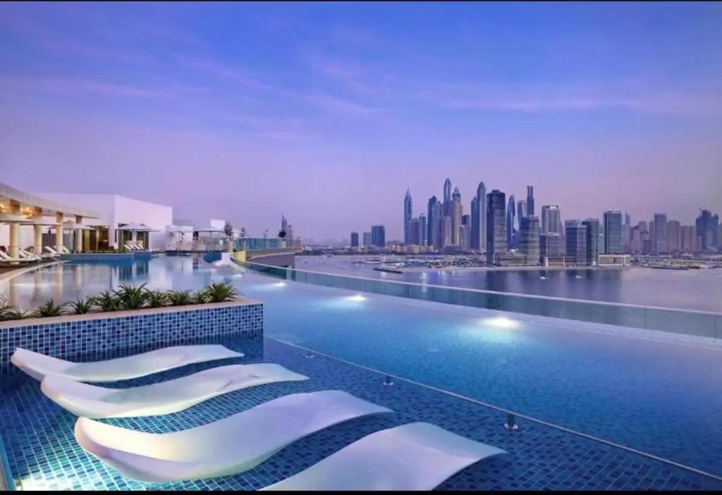 An infinity rooftop pool stretches across the image, reflecting the skyline of Dubai. Lounge chairs line the poolside, offering views of the cityscape. Soft lighting enhances the serene atmosphere, while the horizon blends seamlessly with the sunset hues.