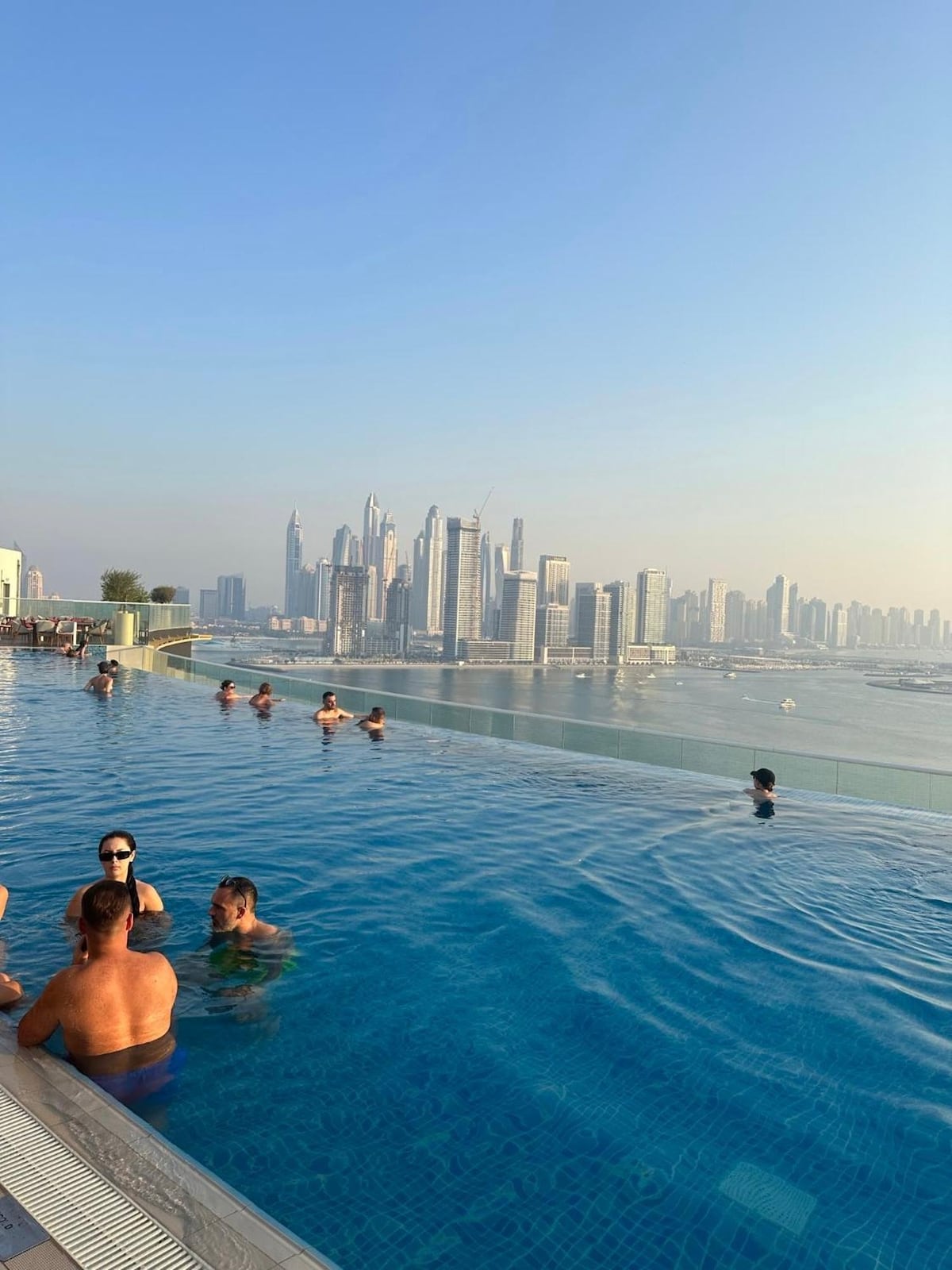 An inviting infinity rooftop pool is visible, offering panoramic views of Dubai's skyline. Several individuals are enjoying the water, with the city's tall buildings and the serene sea in the background. The clear blue sky enhances the tranquil atmosphere surrounding the pool.
