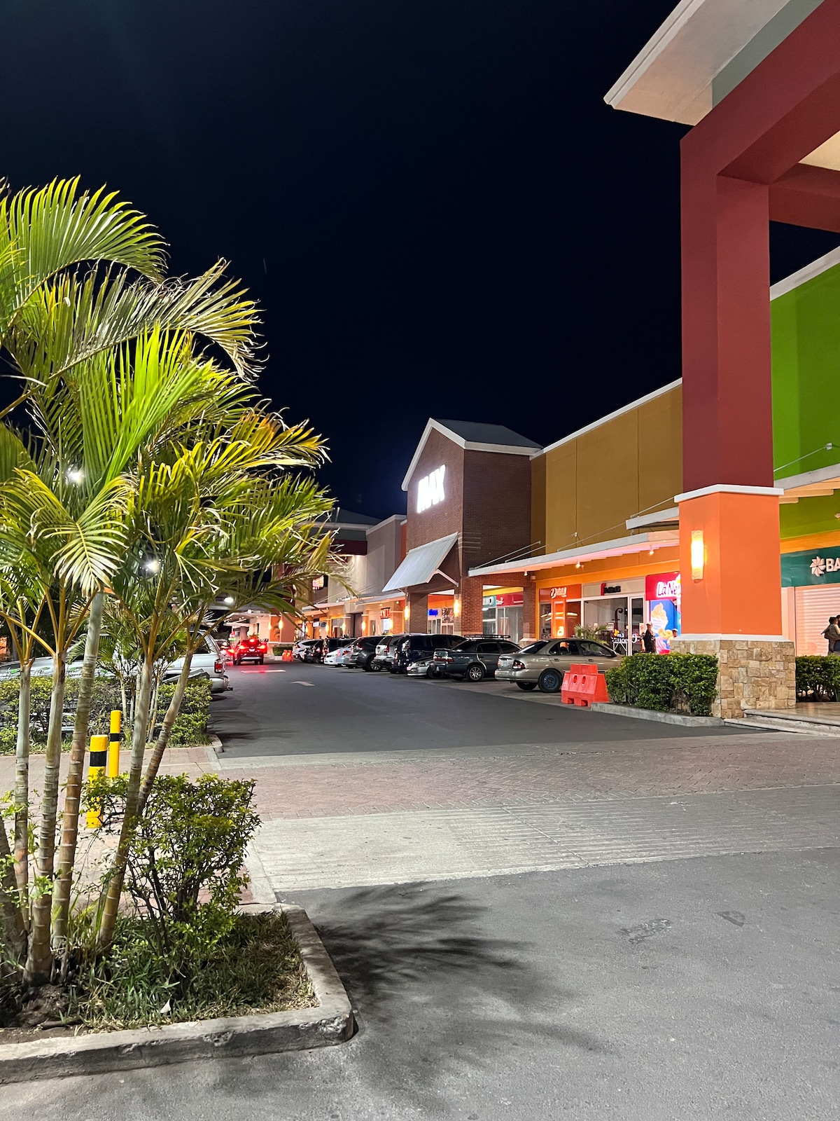 A nighttime view of a parking area lined with palm trees and greenery. The colorful storefronts are illuminated, with parked cars visible along the paved pathway. Soft lighting enhances the inviting atmosphere of the shopping complex.